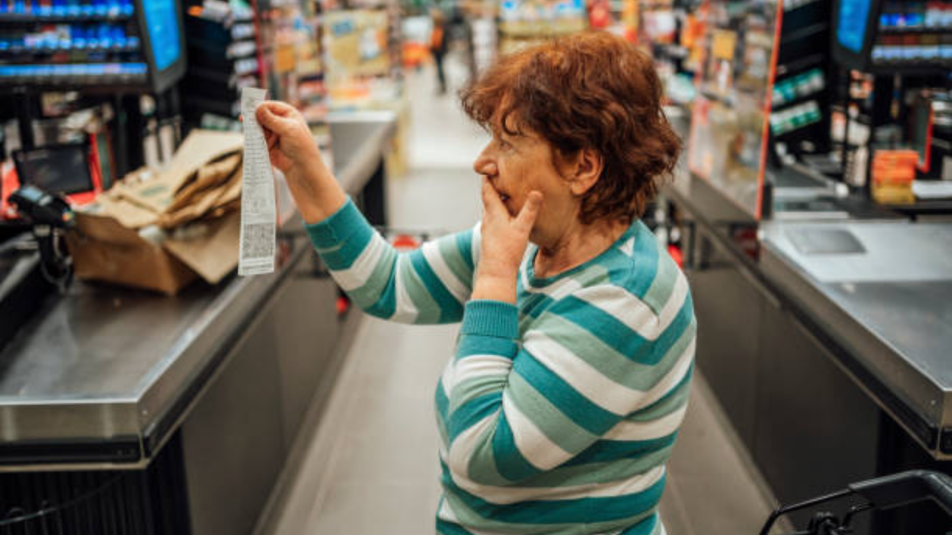 An elderly woman in a striped sweater looking shocked and covering her mouth while reviewing a long grocery receipt.