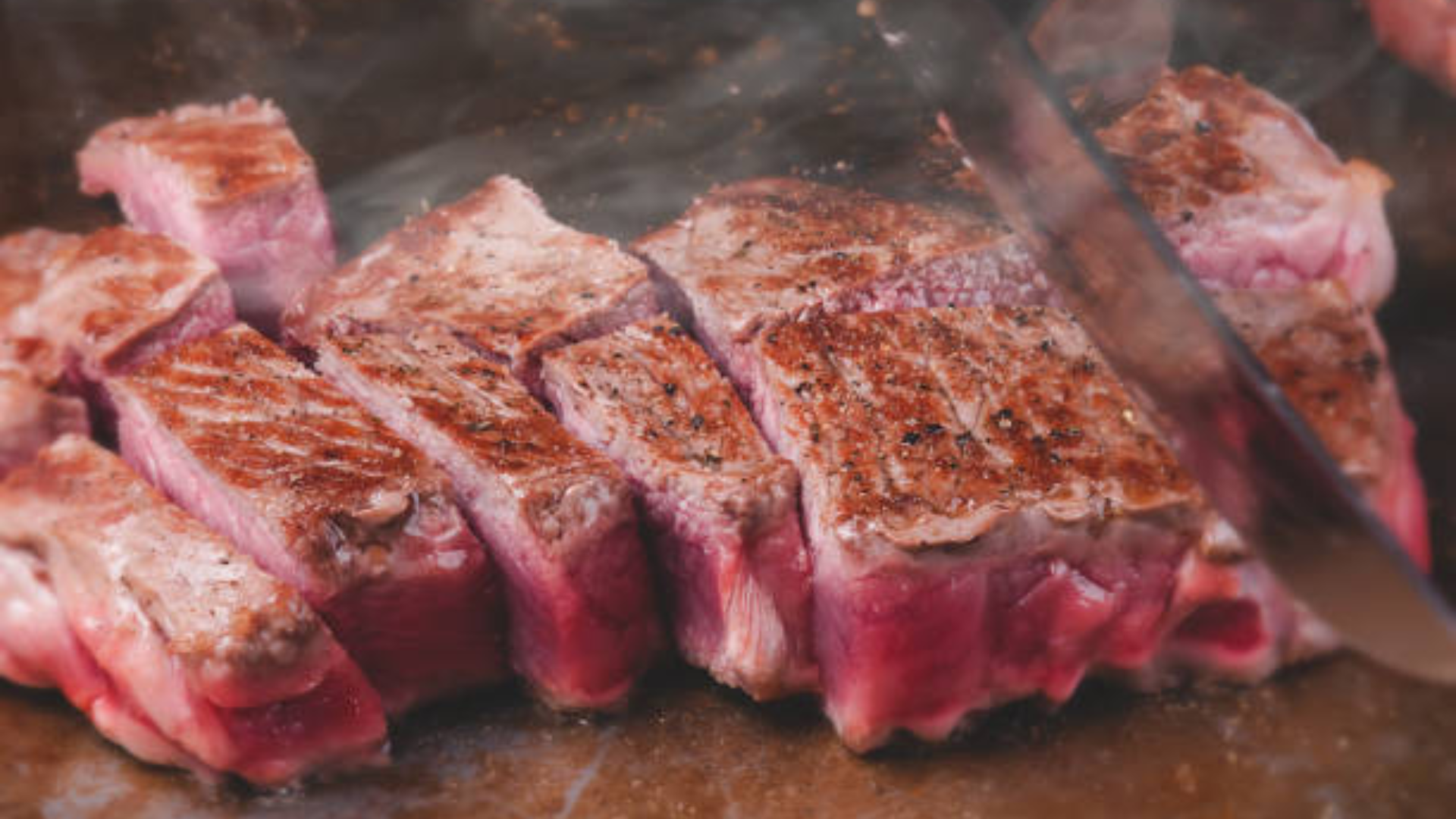 Close-up of thick steaks being sliced on a hot grill with steam and char visible on the meat.