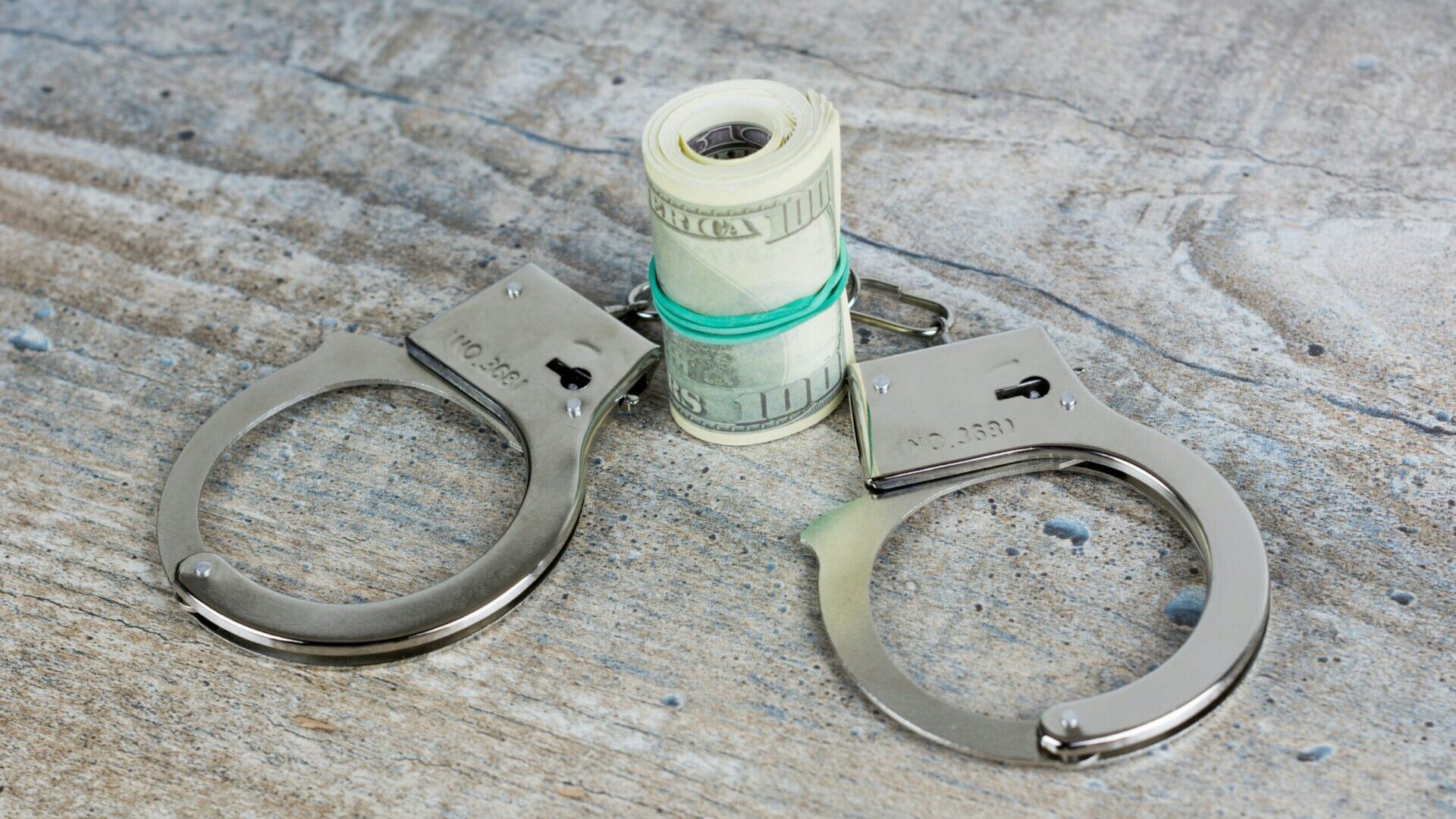 Scissors cutting through a rolled stack of U.S. dollar bills on a table.