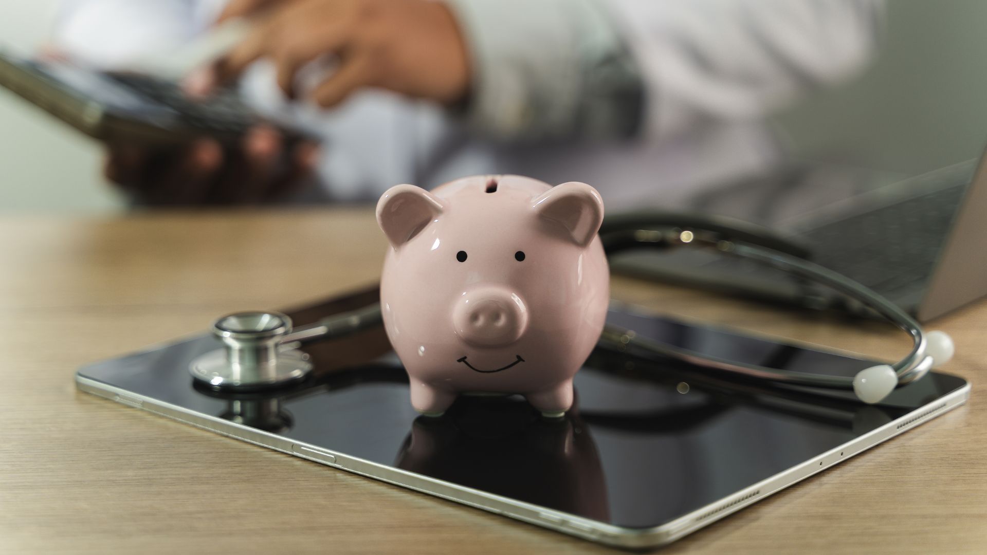 Pink piggy bank sitting on a tablet with a stethoscope and phone on a desk.