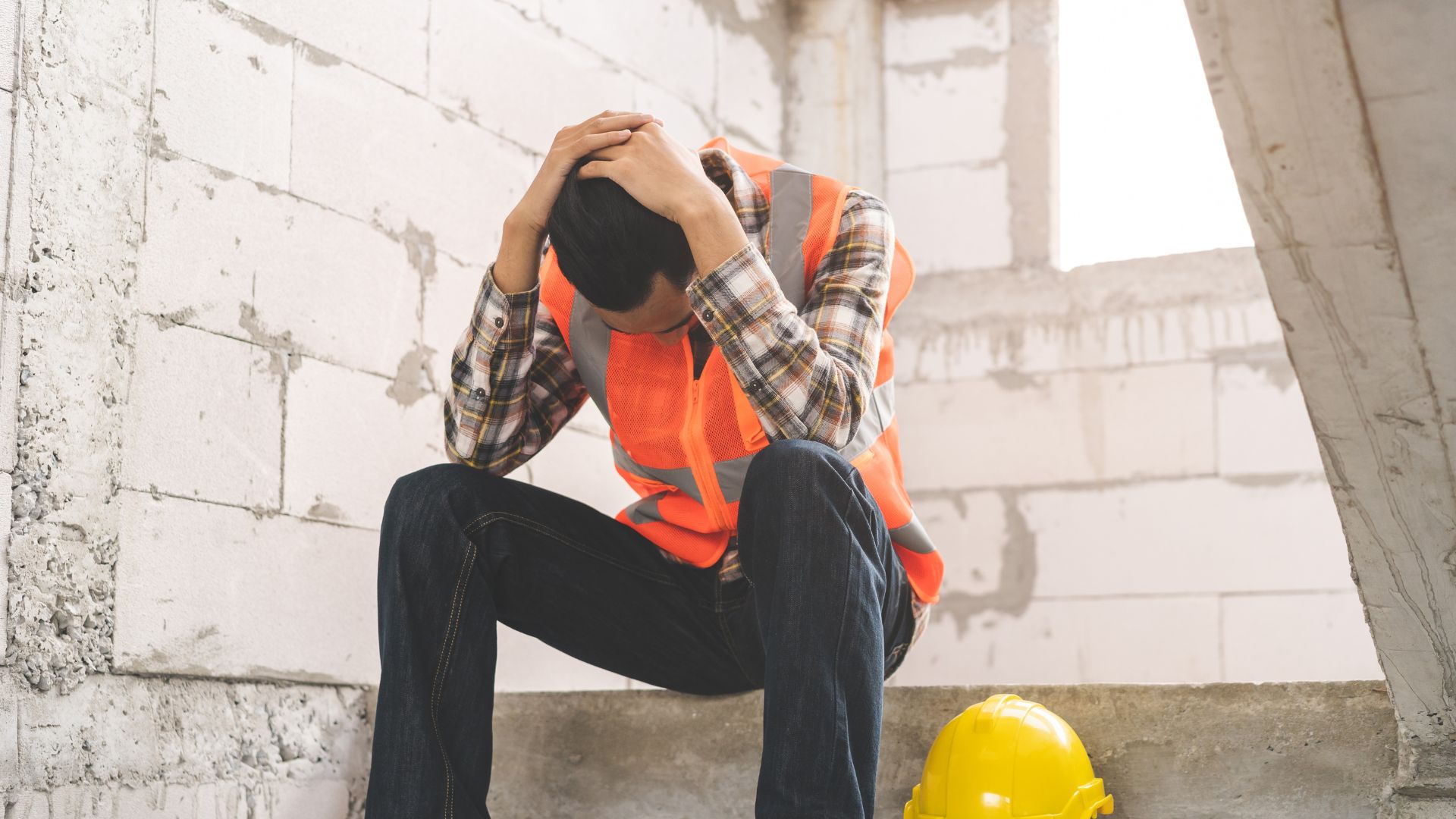 Construction worker in a safety vest sitting with his head in his hands next to a yellow hard hat.
