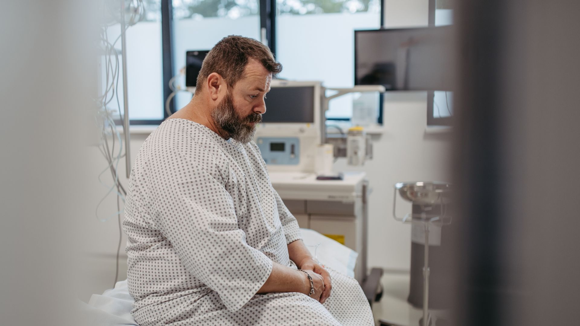 Hospital patient wearing a gown sitting on a bed in a medical room.