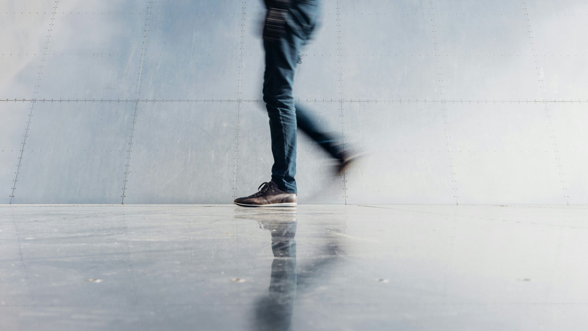 Person walking across polished floor with reflection against a metallic wall background.