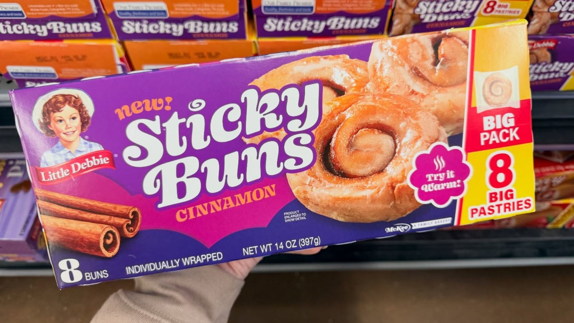A hand holding a box of Little Debbie's new BIG PACK Sticky Buns with other boxes at the back in a Walmart.