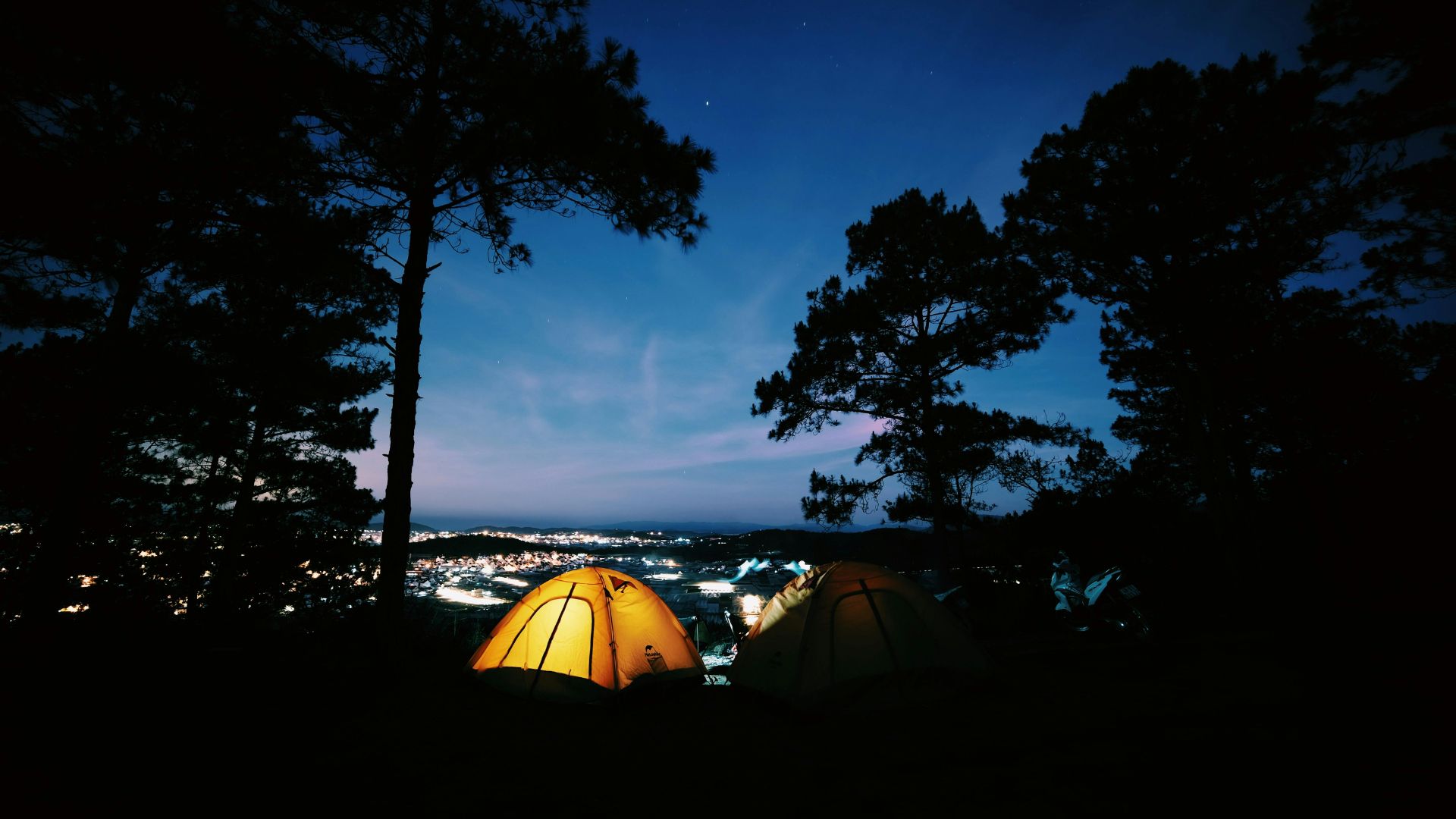 Glowing orange tent at a wooded campsite at night with a starlit sky and city lights in the distance.