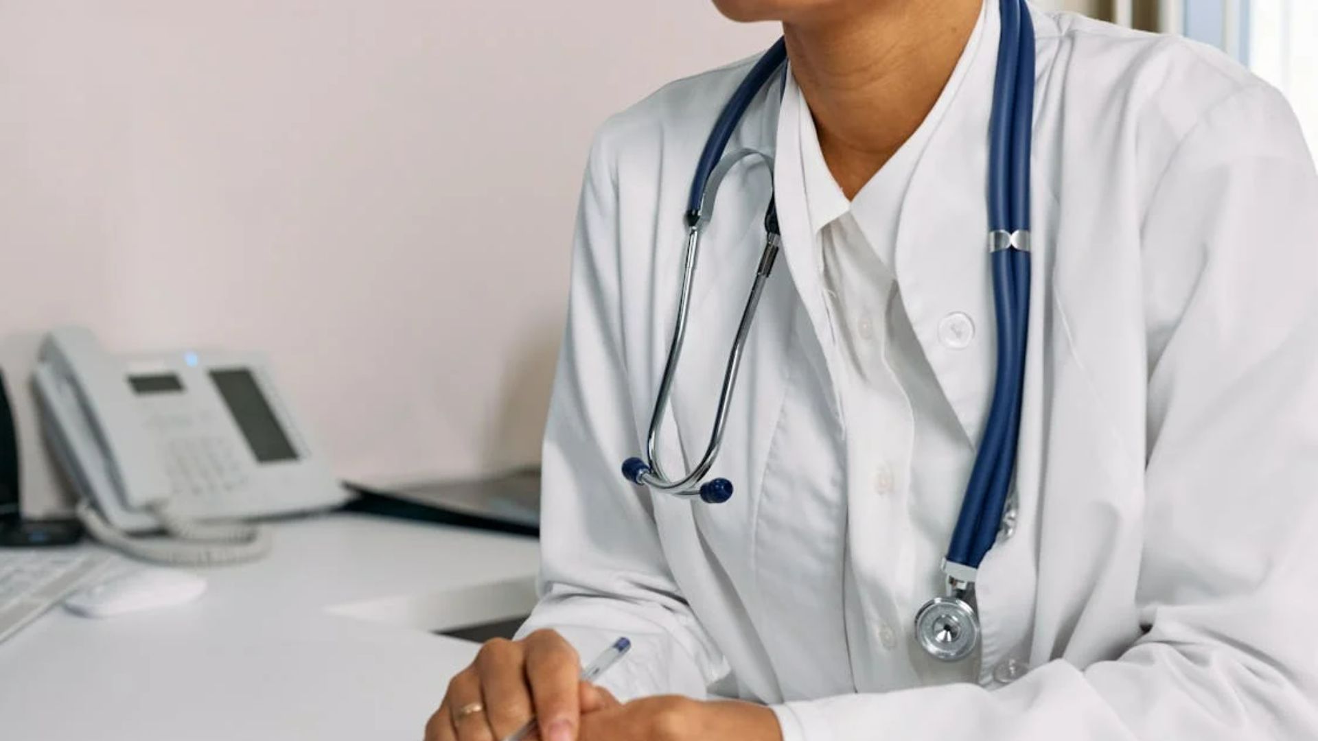 A doctor in a white coat with a stethoscope around their neck, seated at a desk holding a pen.