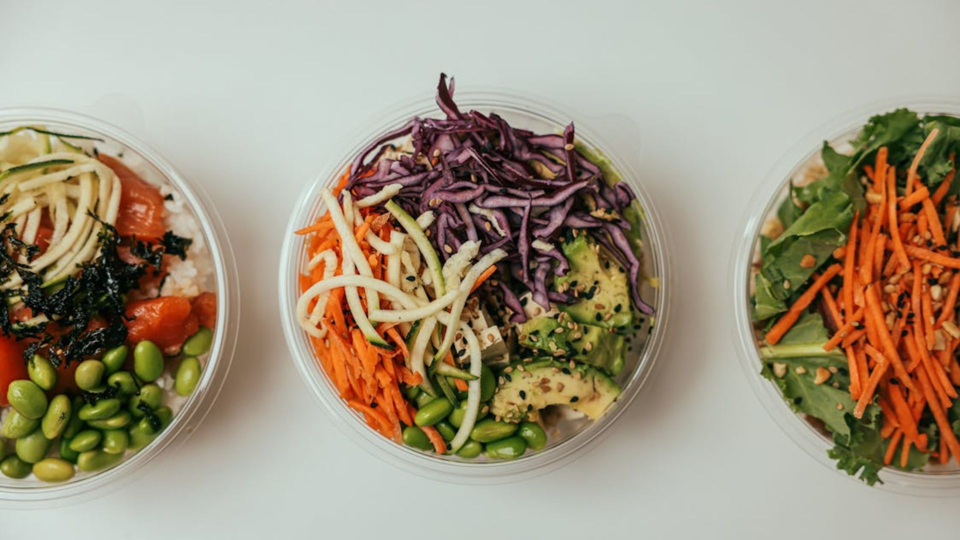 Three colorful poke bowls in clear containers arranged in a row, topped with ingredients like avocado, shredded cabbage, carrots, edamame, seaweed, and rice.