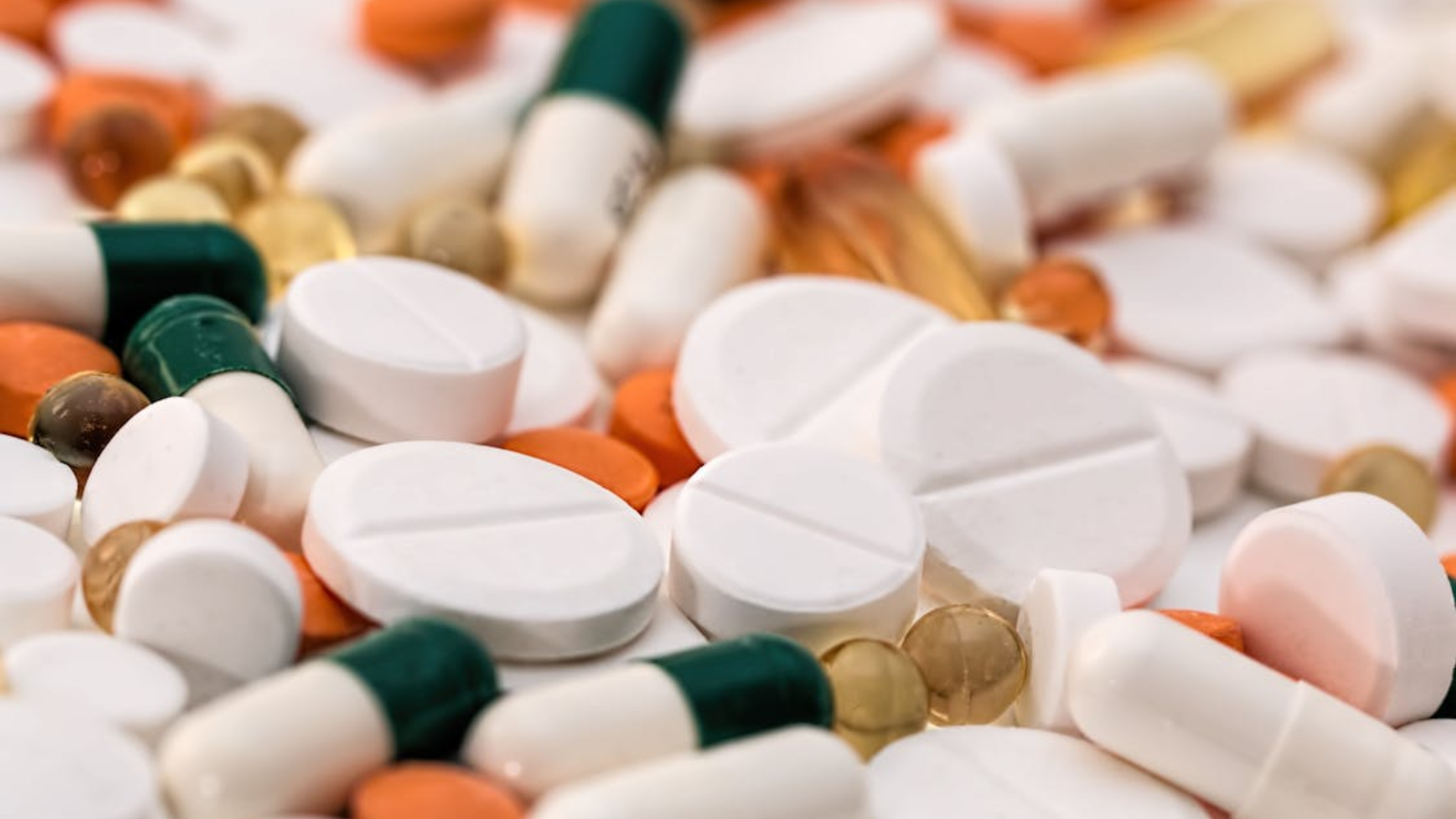A scattered pile of various medications and supplements, including white circular tablets, green and white capsules, and orange pills.