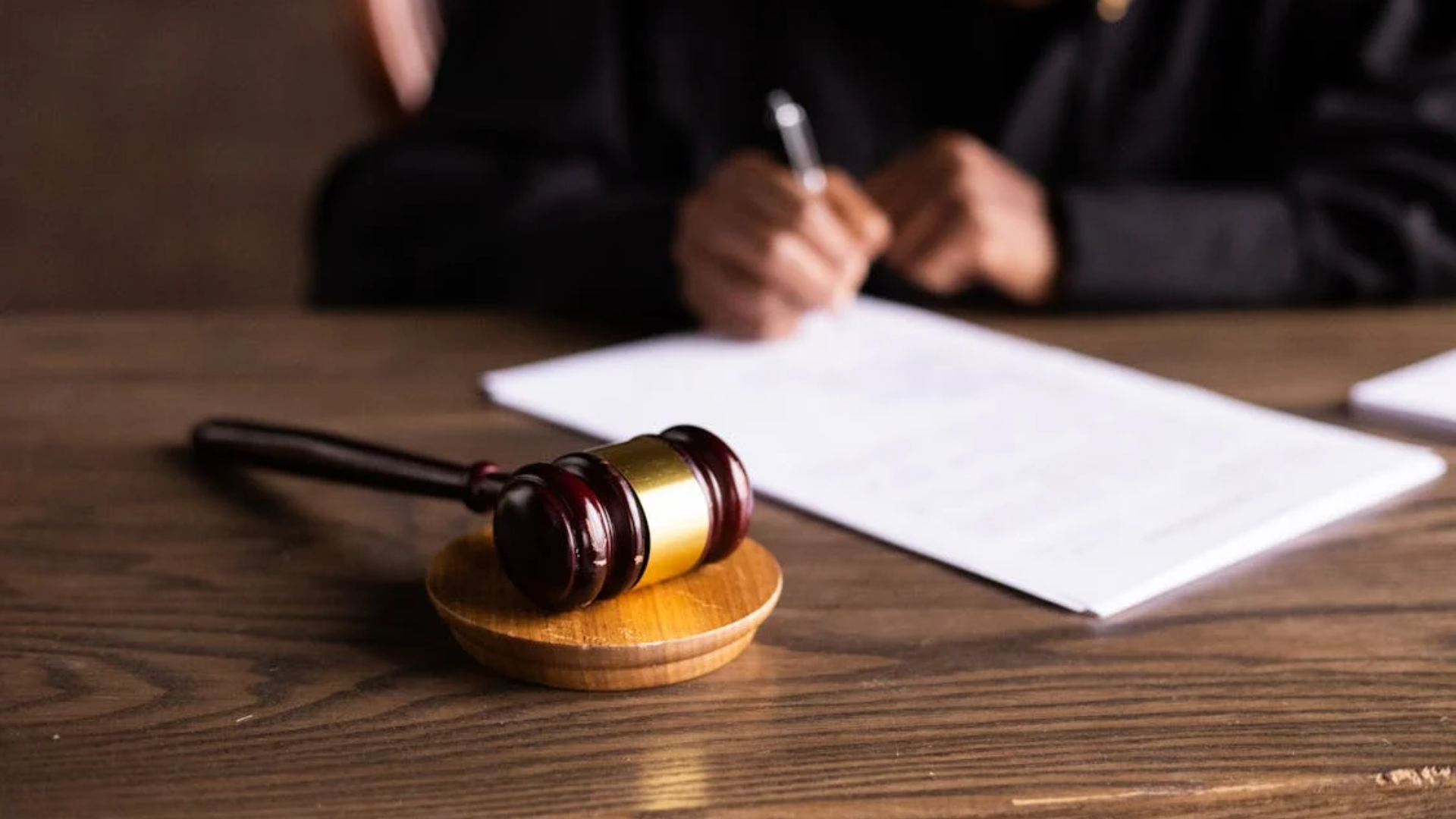 A wooden gavel and sound block rest on a desk in the foreground, with a judge in black robes writing on a document in the blurred background.