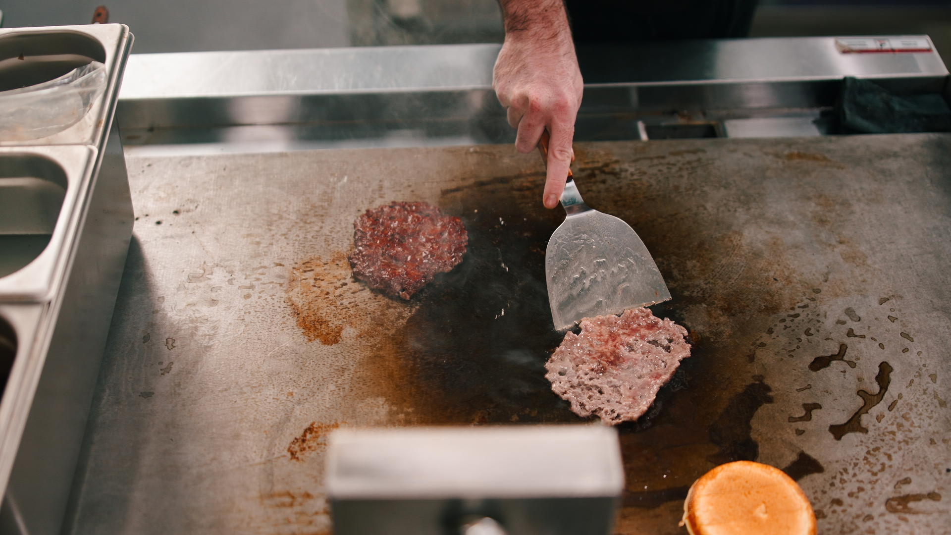 A close-up of a cook's hand using a metal spatula to press down and sear a thin hamburger patty on a hot flat-top grill.