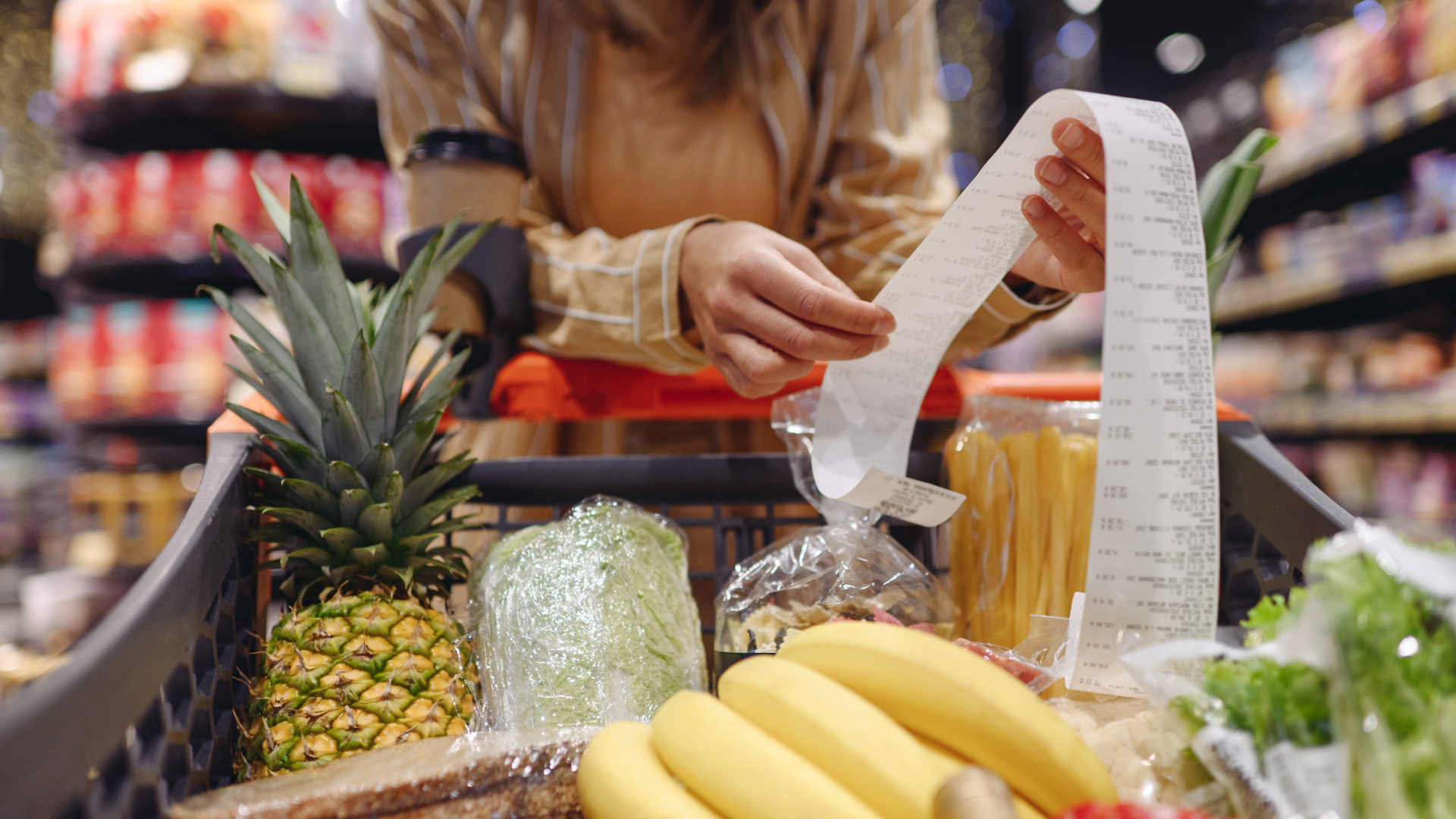 A person looking at a long receipt while leaning over a shopping cart full of fresh produce like pineapple and bananas.