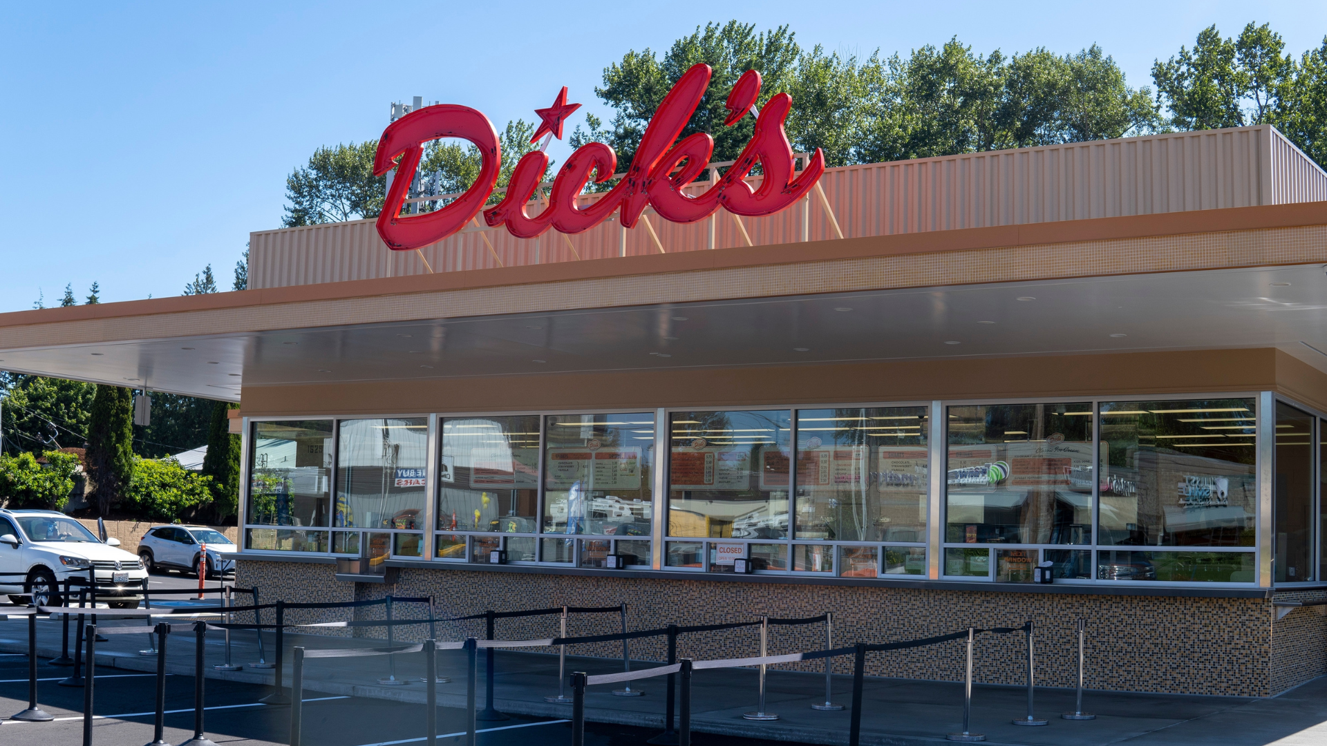 The exterior of a _Dick's Drive-In_ restaurant, showing the service windows, outdoor queue area, and iconic red neon sign against a clear blue sky.