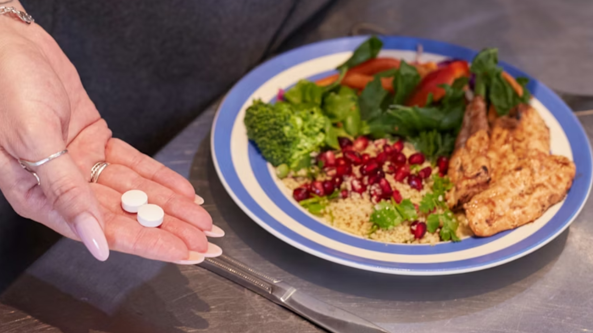 A person holding two white tablets in one hand and a glass of water with a lemon slice in the other, standing next to a plate of healthy food including chicken, broccoli, and pomegranate.
