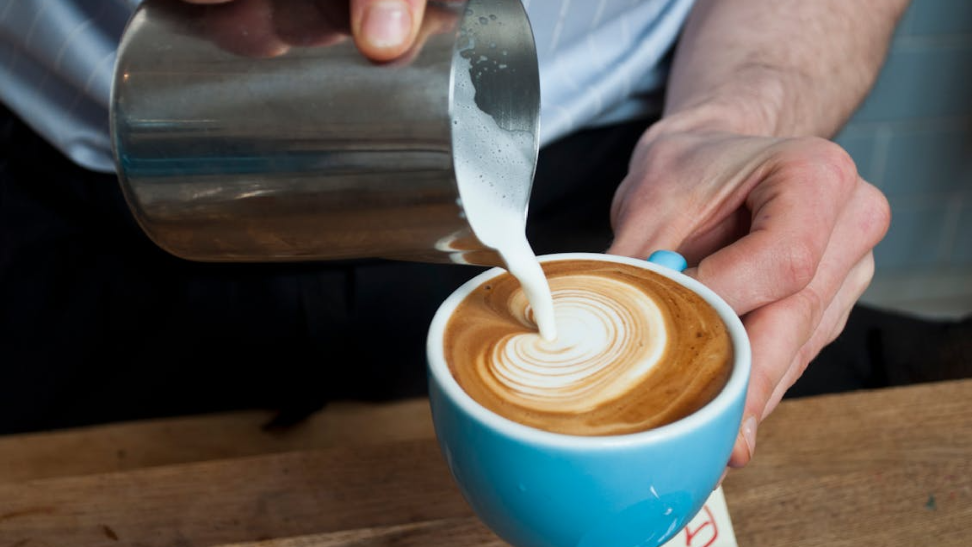 A person pouring steamed milk from a metal pitcher into a blue cup to create latte art.