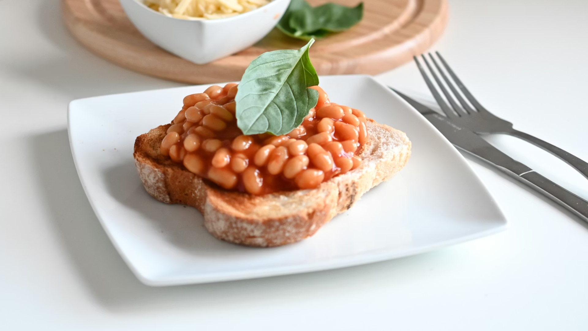 A single slice of thick, toasted sourdough bread topped with a heap of baked beans and a fresh basil leaf, served on a white square plate with a side of shredded cheese.