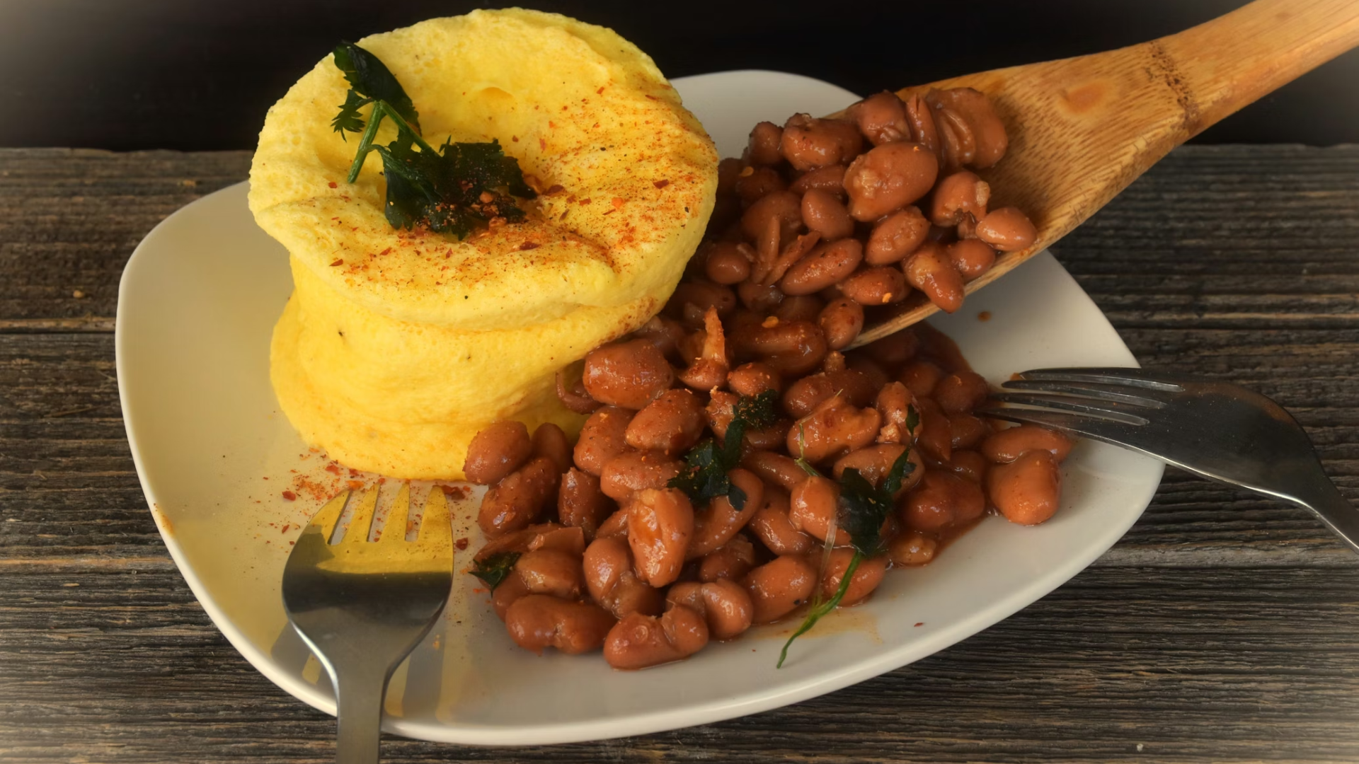 A white plate featuring a large, fluffy yellow omelet topped with herbs, served alongside a pile of savory brown beans.