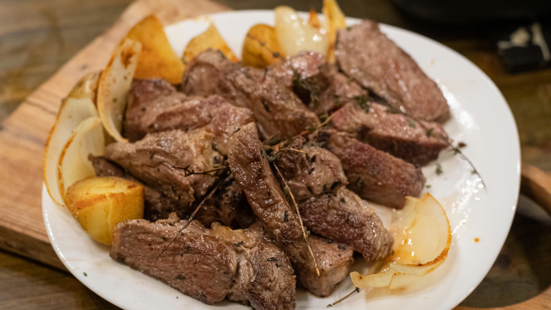 A white plate filled with sliced, cooked steak seasoned with herbs, served with roasted potatoes and onions.