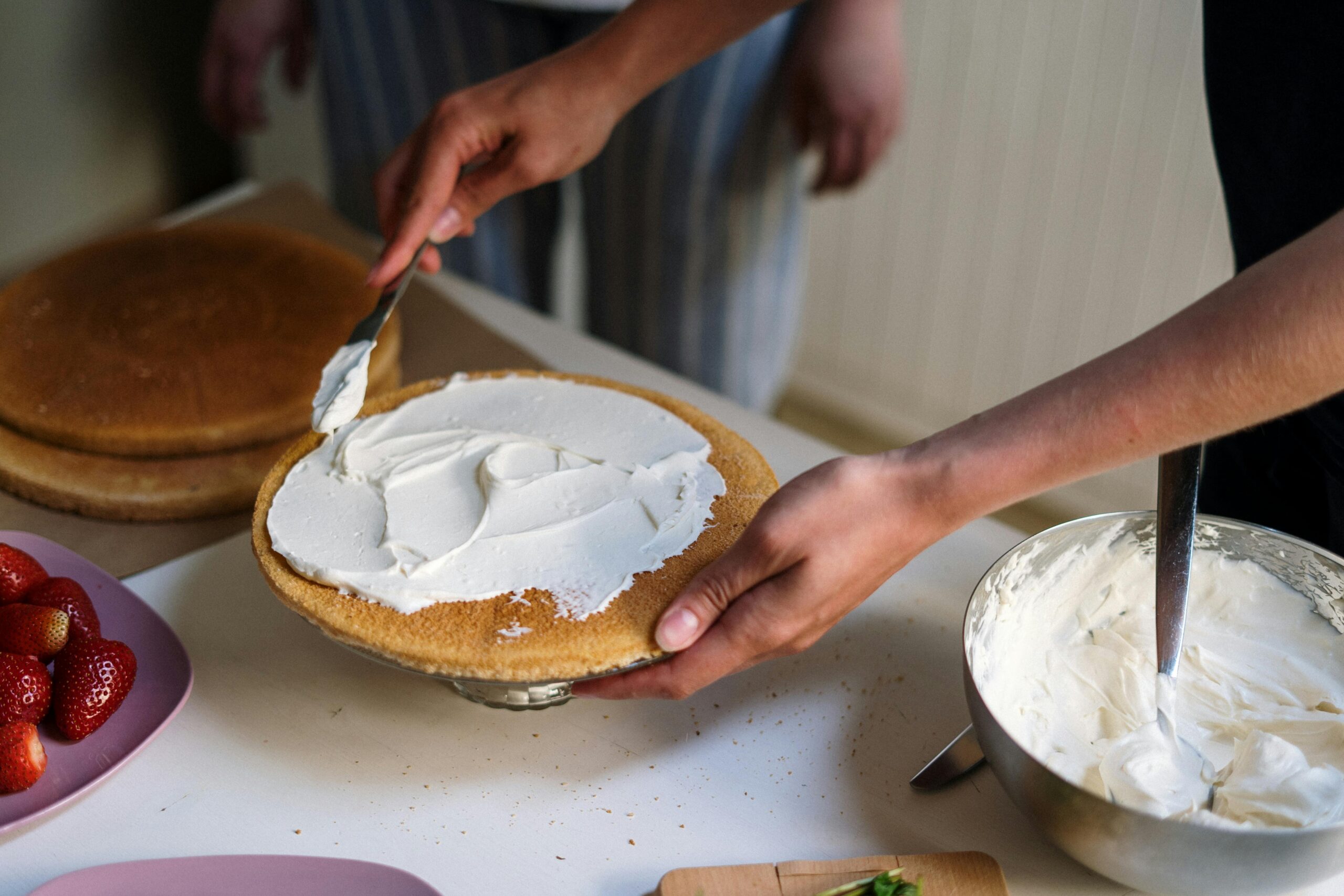 Person spreading white frosting onto a round cake layer on a kitchen counter.