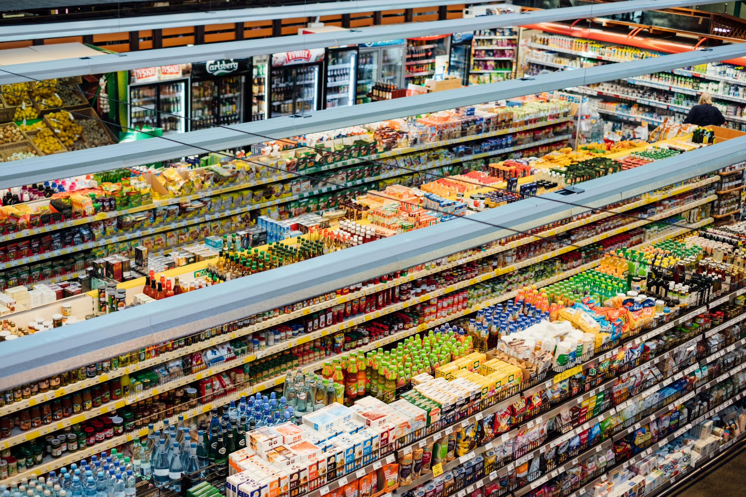 Wide view of a supermarket aisle stocked with packaged foods, drinks, and refrigerated items on shelves.