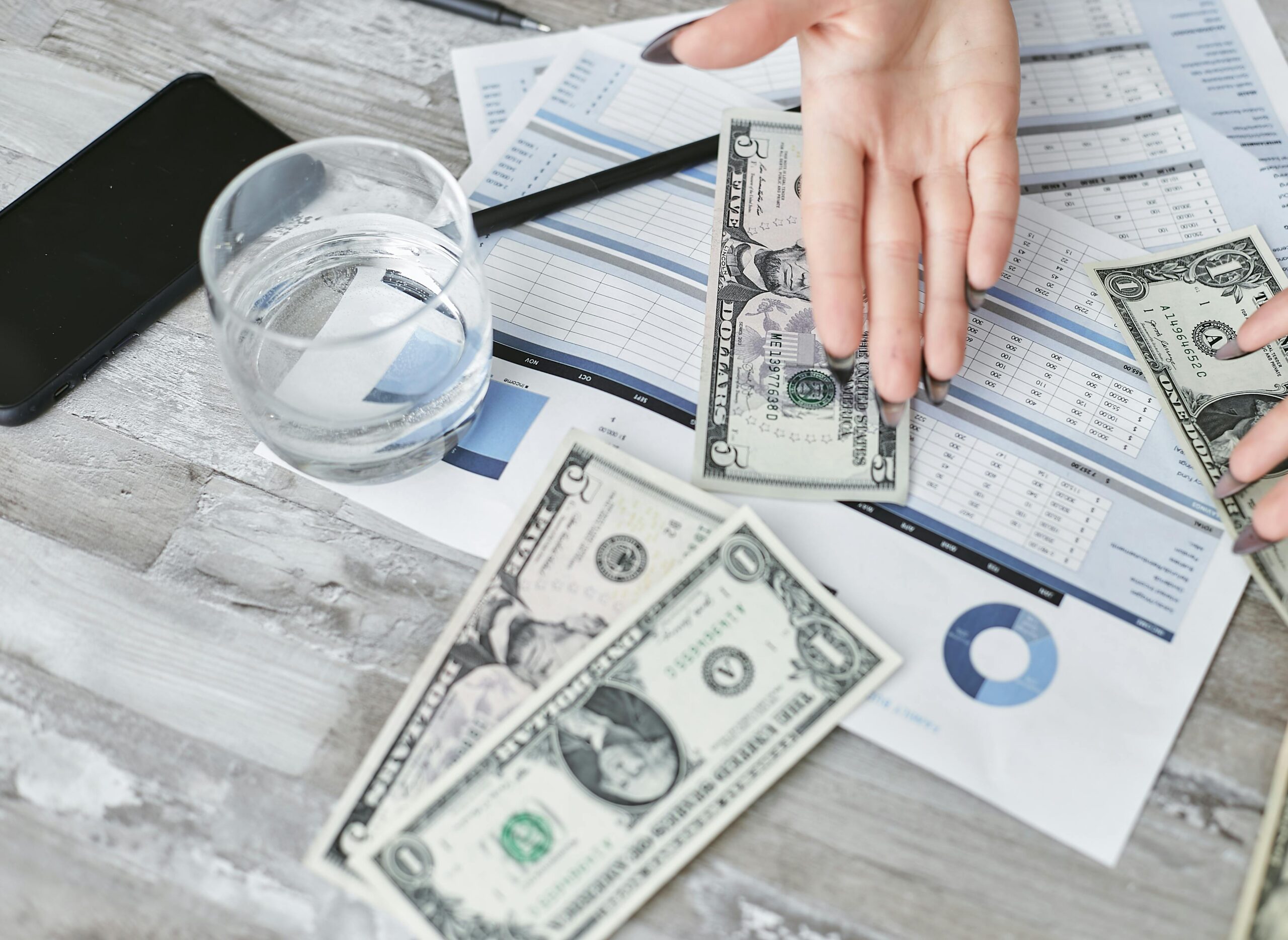 Hand placing dollar bills on financial documents with charts and tables on a desk.