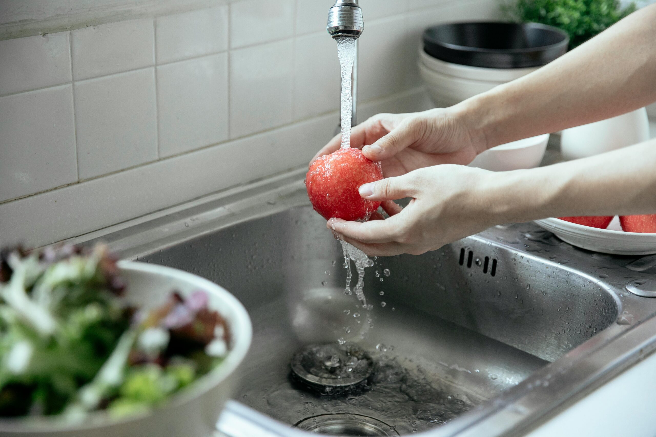 Hands rinsing a red apple under running kitchen faucet.