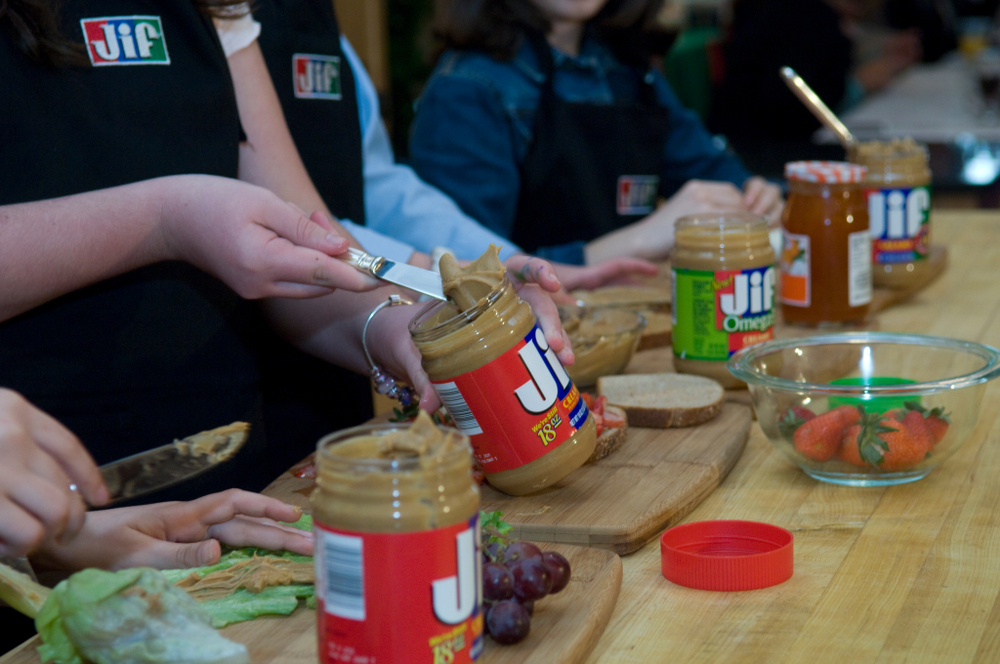 Children spreading Jif peanut butter onto bread at a table with fruit and jars during a food prep activity.