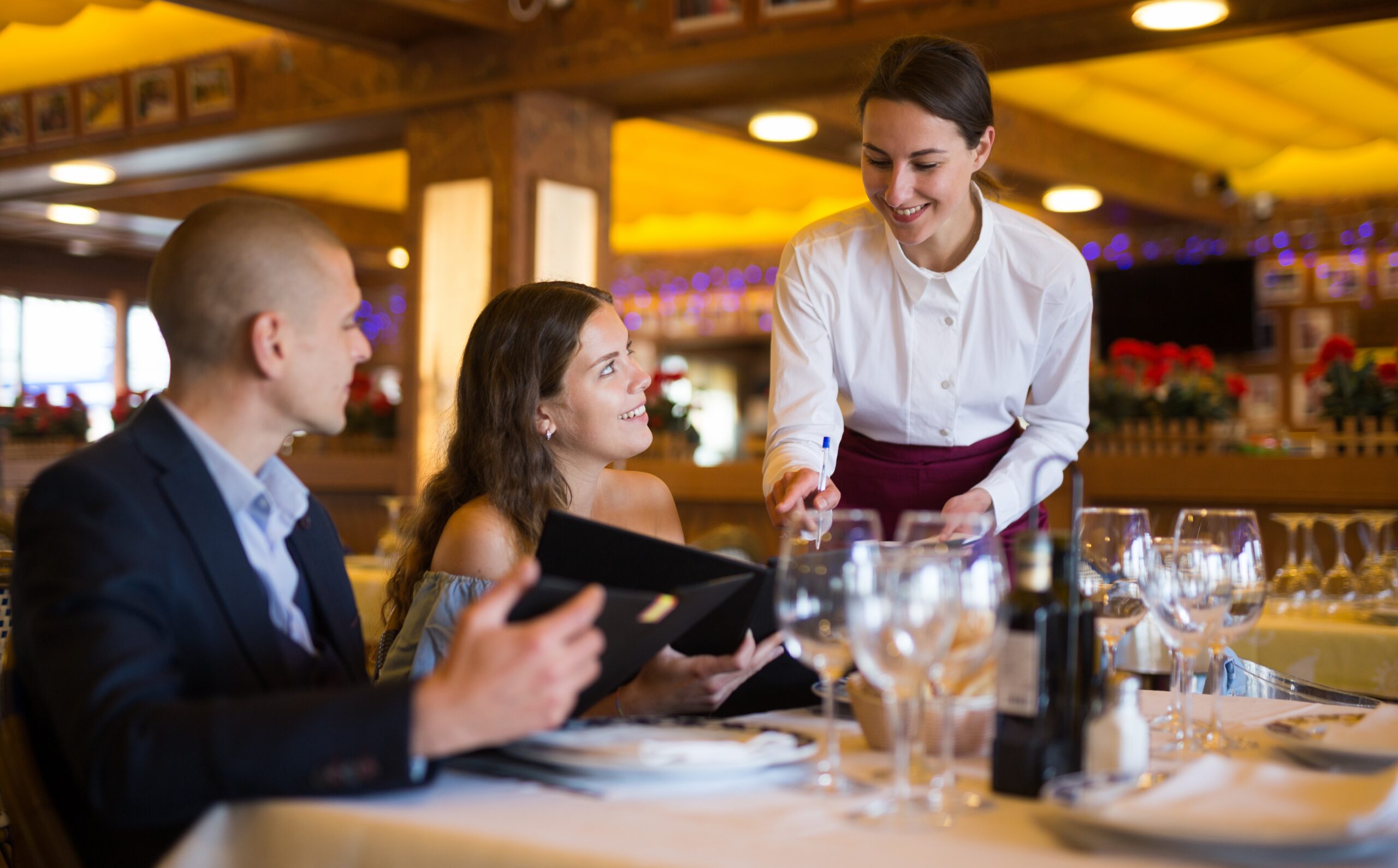 Server taking an order from a couple seated at a restaurant table with menus and wine glasses.