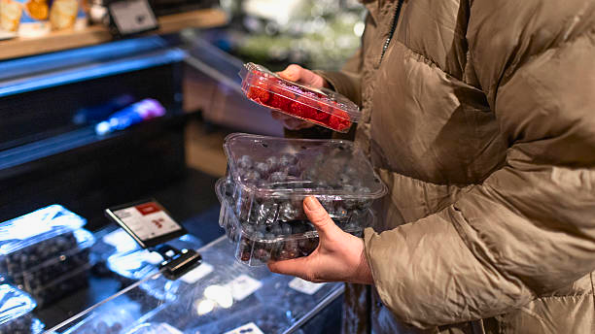 A close-up of a person in a tan puffer jacket holding several plastic containers of fresh blueberries and raspberries in a grocery store produce section.