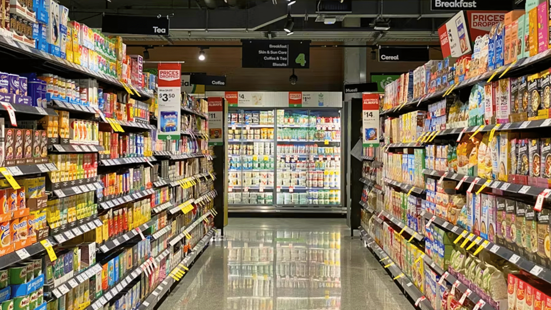 A wide, high-angle shot looking down a brightly lit supermarket aisle with polished grey floors; shelves on both sides are fully stocked with colorful boxed and canned goods.