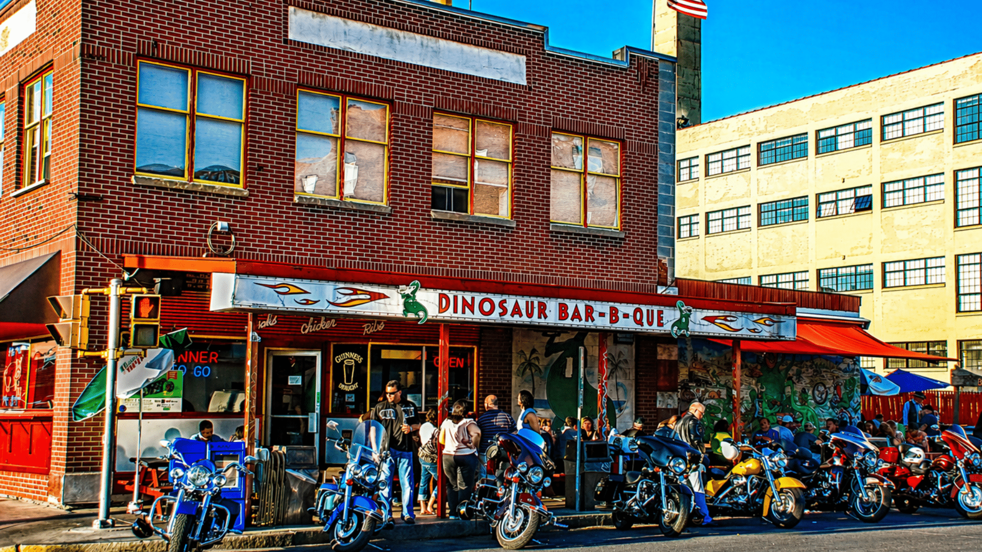 A high-contrast daytime shot of the original Syracuse Dinosaur Bar-B-Que brick building; several motorcycles are parked along the sidewalk in front of the red-and-white awning, and a crowd is gathered near the entrance under a clear blue sky.