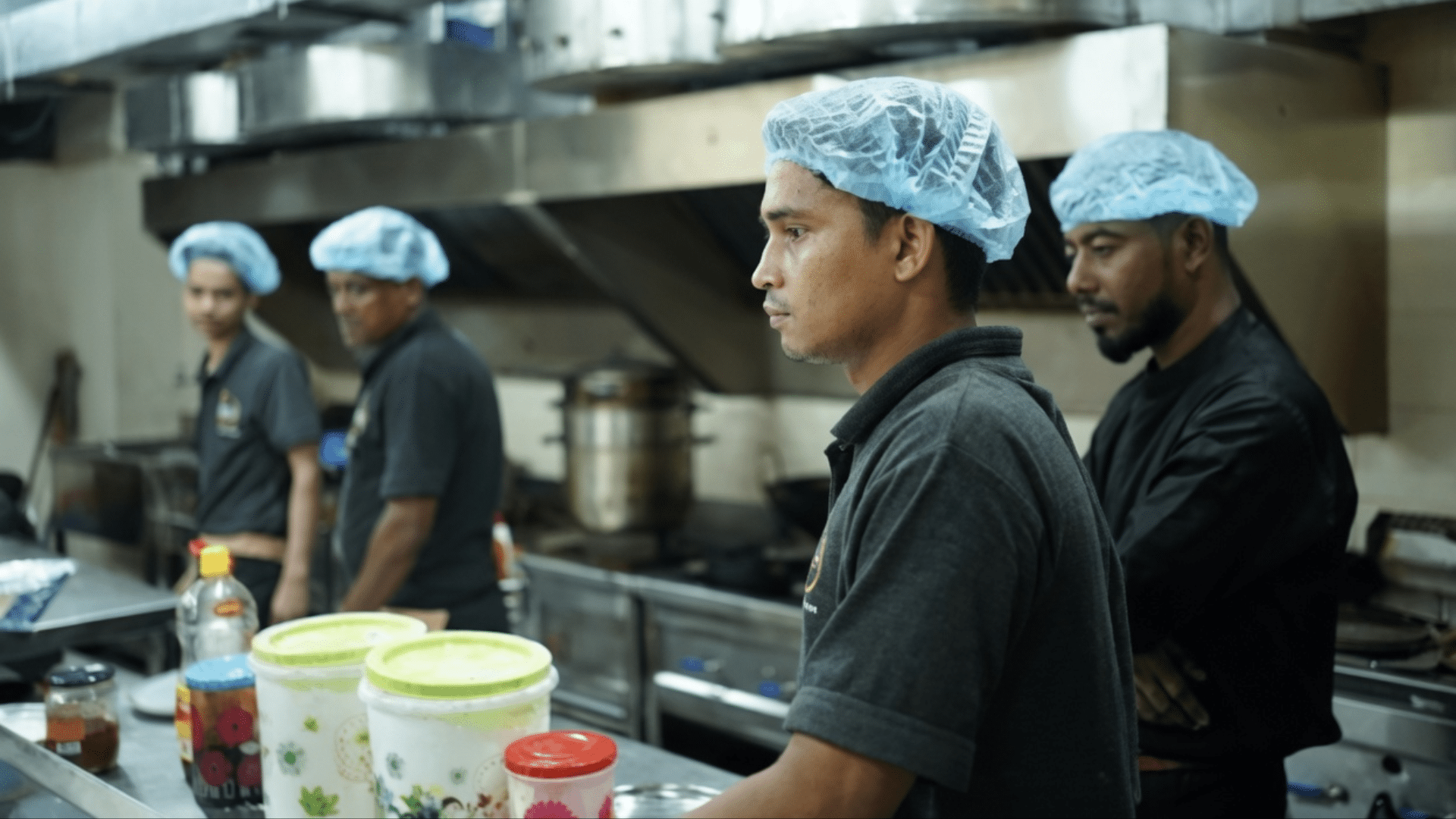 A line of male kitchen staff wearing blue hairnets and black polo shirts standing in a stainless-steel commercial kitchen.