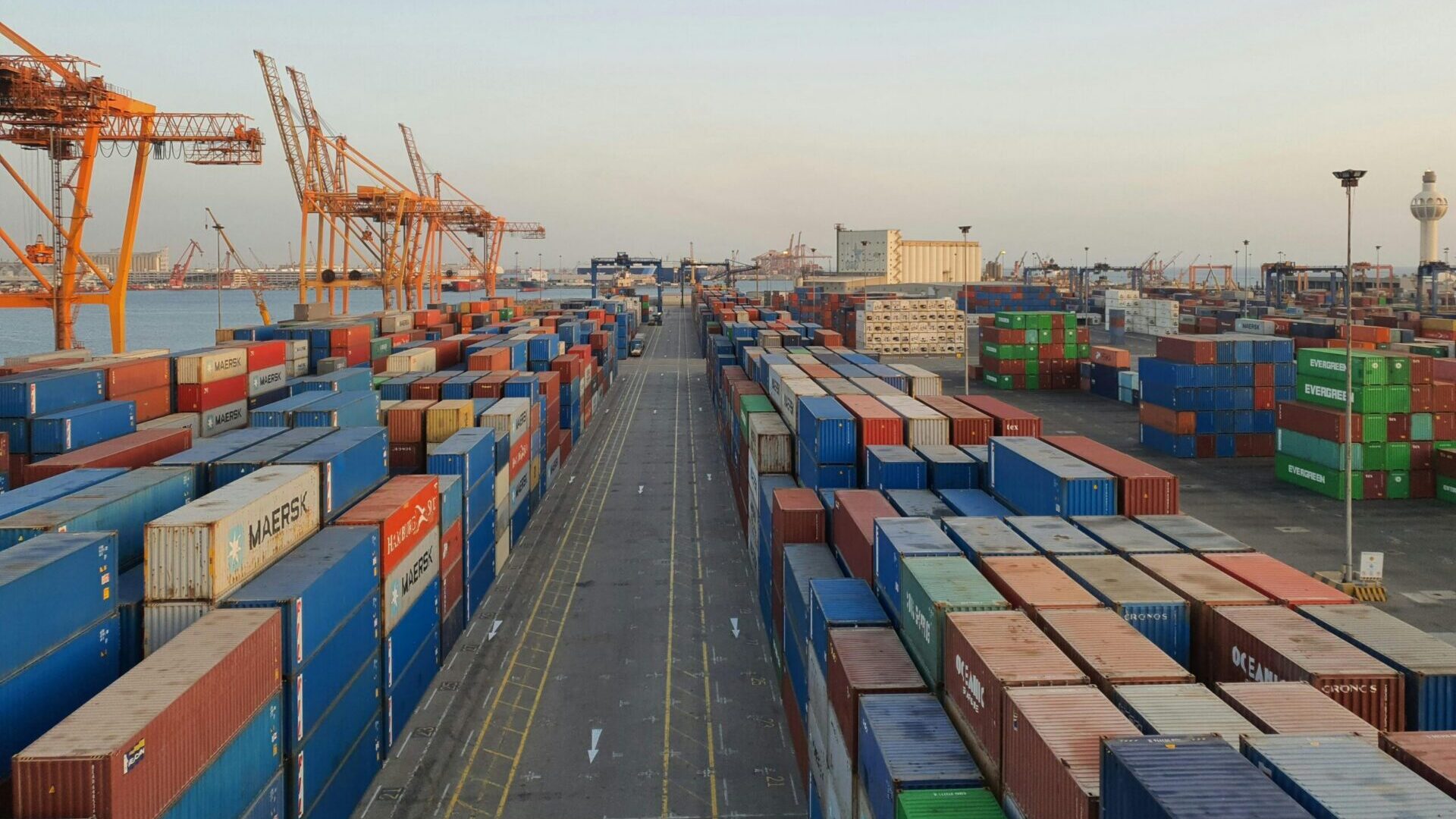 Shipping containers stacked in rows at a busy port with cranes in the background.