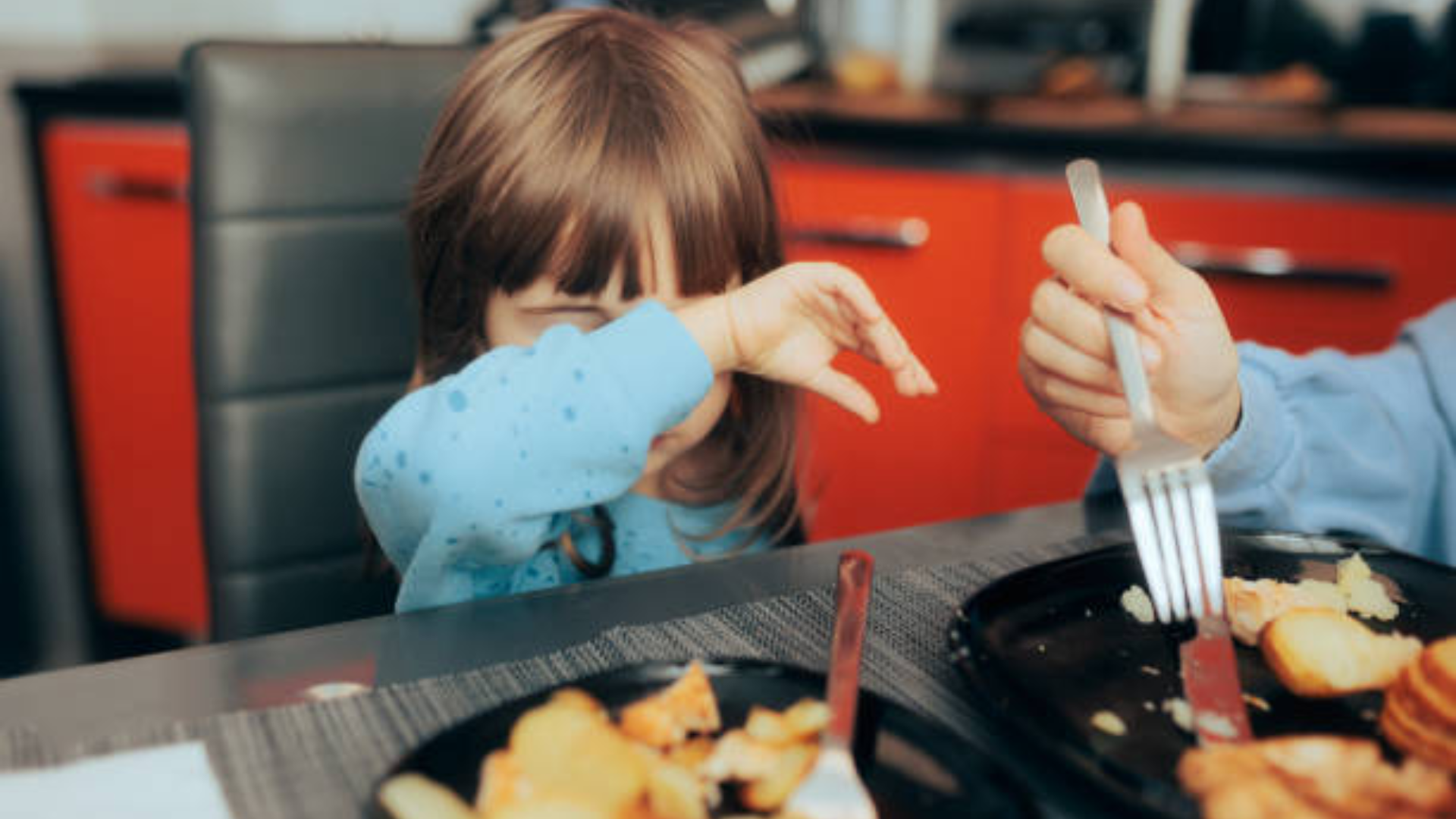 A young girl at a dinner table, crying and wiping her eyes with her arm while someone next to her eats.