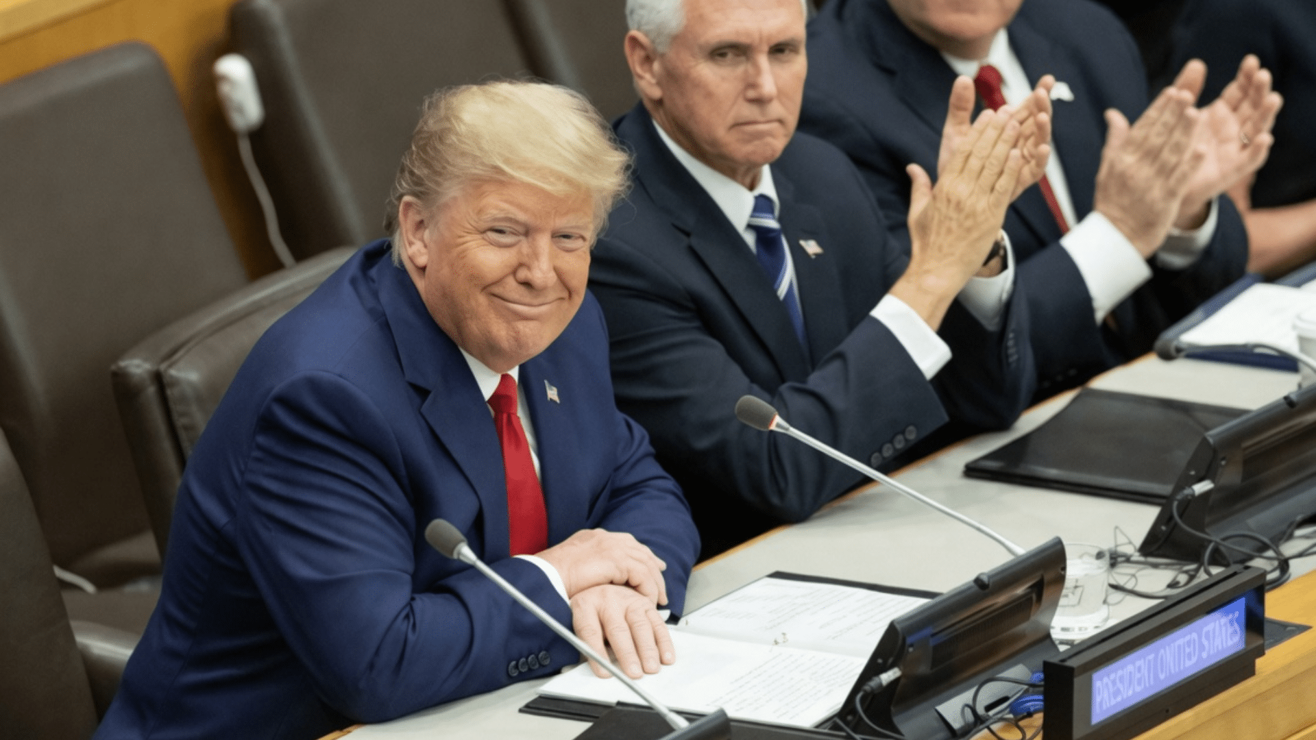 A medium shot of President Donald Trump sitting at a formal conference table, smiling and looking toward the camera.