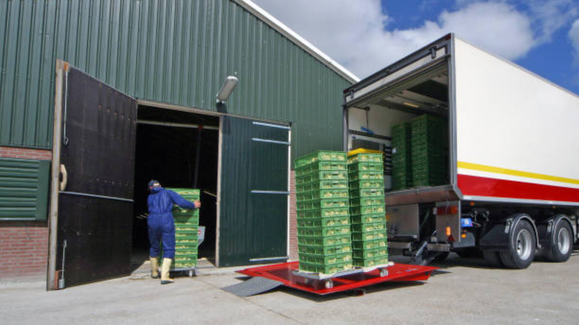 A worker in blue overalls loading tall stacks of green plastic crates into the back of a refrigerated semi-truck outside a large warehouse.