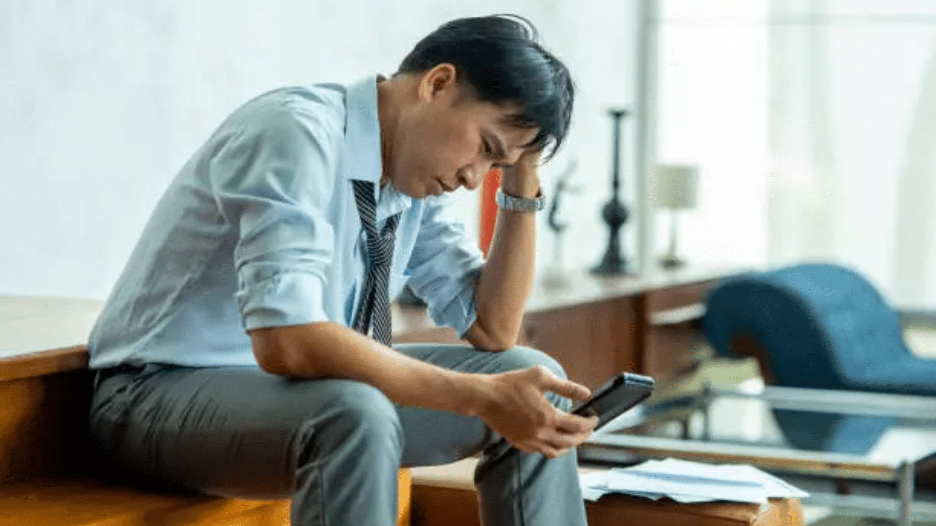A medium shot of a businessman in a light blue shirt and tie sitting on wooden stairs, looking dejected and resting his head in his hand while staring at his smartphone, reflecting stress.