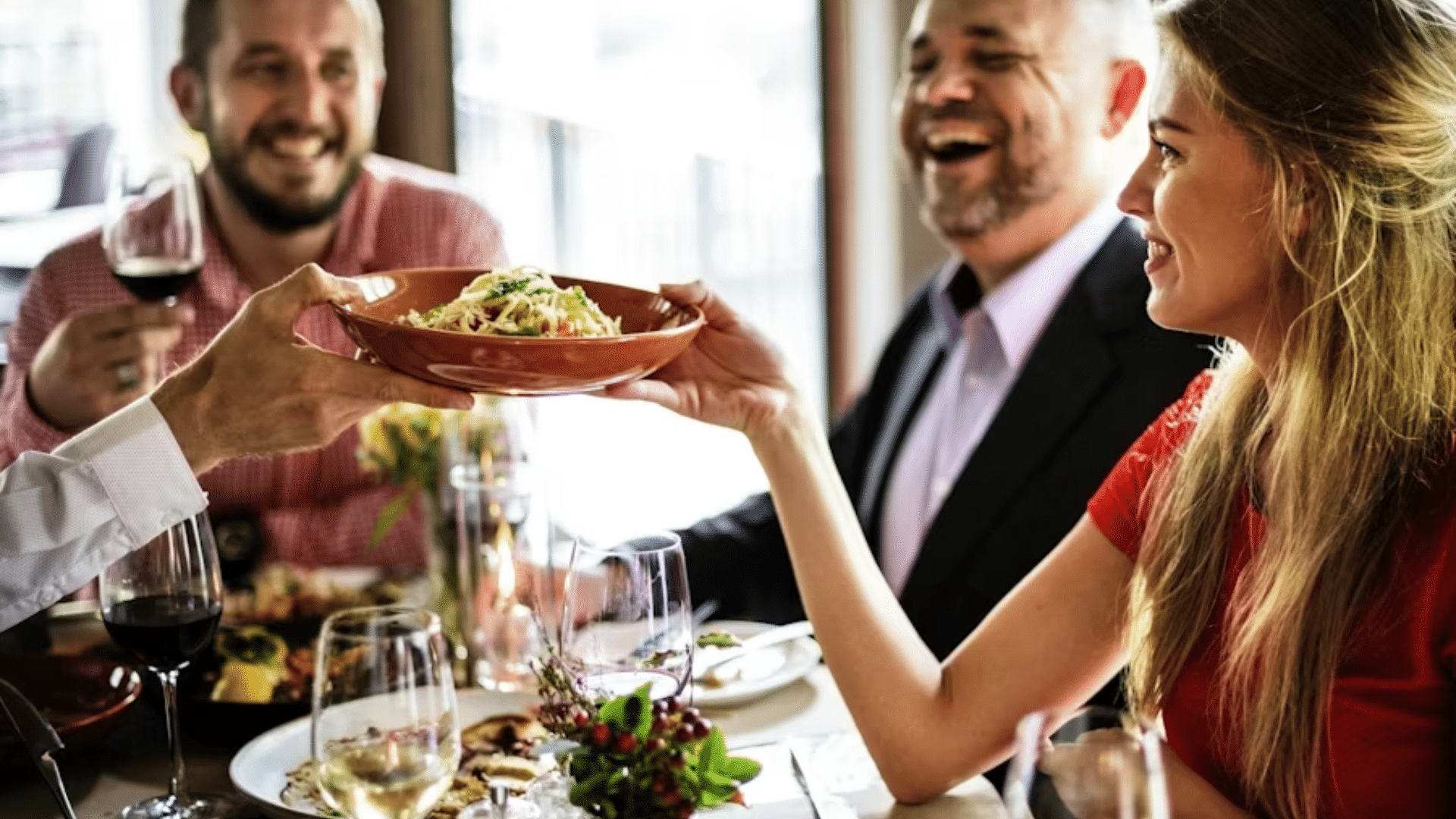 A close-up shot of a joyous dinner party where a hand is reaching across the table to share a bowl of pasta with a woman in a red dress, while men in the background laugh and enjoy wine.