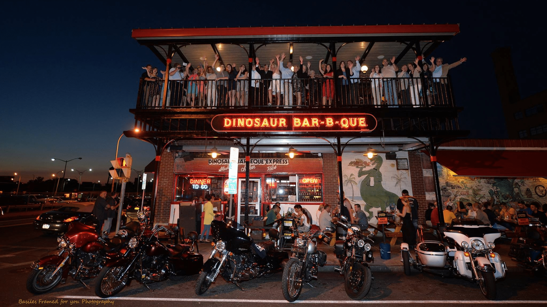 A vibrant nighttime shot of the Brooklyn Dinosaur Bar-B-Que on Union Street; a large group of people is waving from the second-story balcony under a glowing neon sign, while a row of motorcycles lines the street in the foreground.