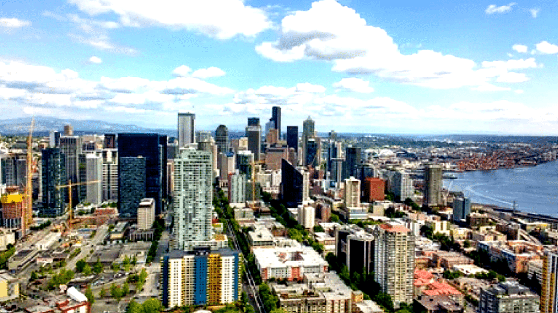 A high-angle aerial view of the Seattle skyline on a bright, partly cloudy day; the dense cluster of glass skyscrapers in the downtown core is visible alongside the deep blue waters of Elliott Bay and the industrial port.