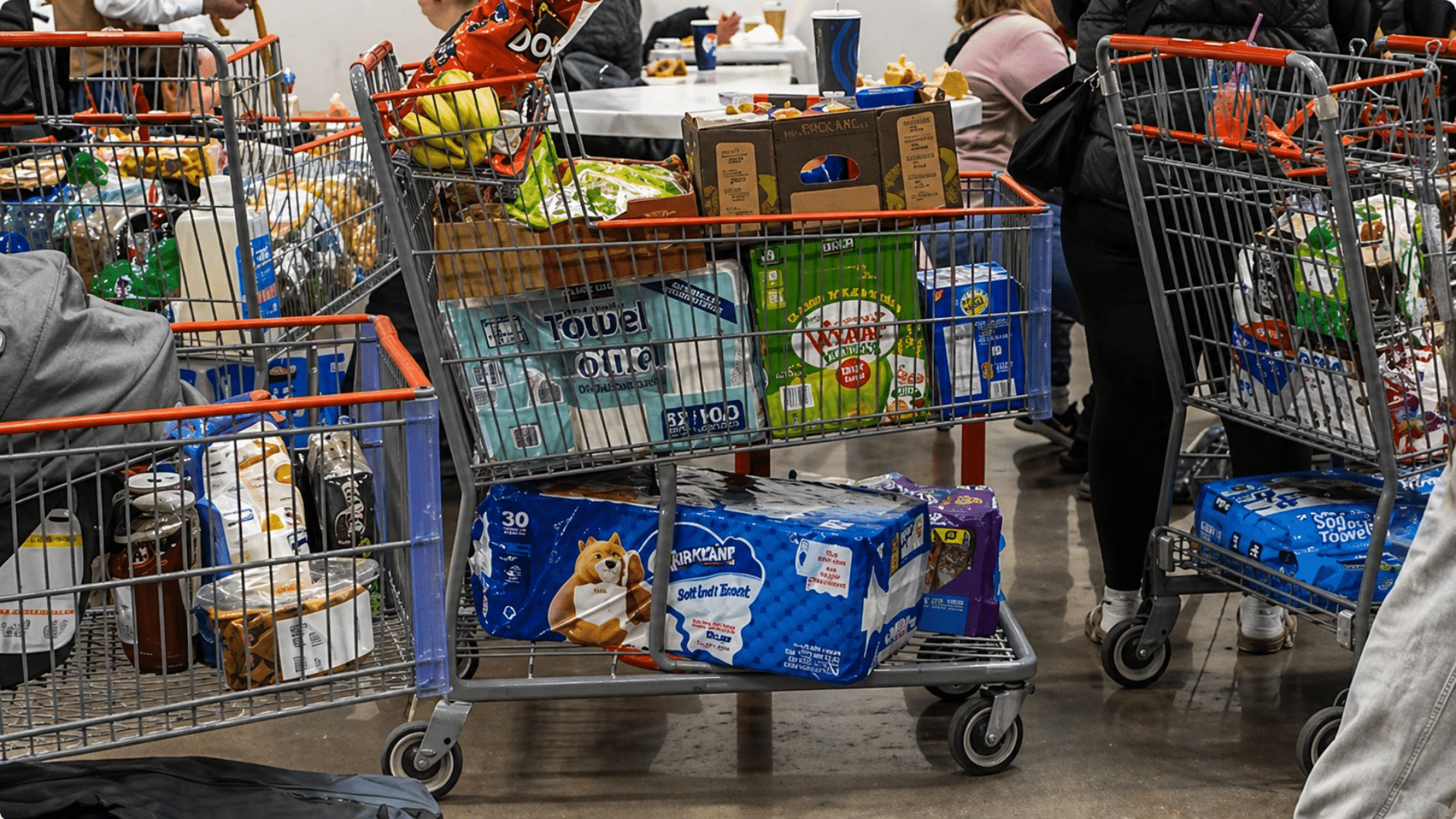 A high-angle shot of several Costco shopping carts crowded together in a warehouse dining area; the carts are filled with bulk items like Kirkland Signature bath tissue, paper towels, large bags of snacks, and fresh produce.
