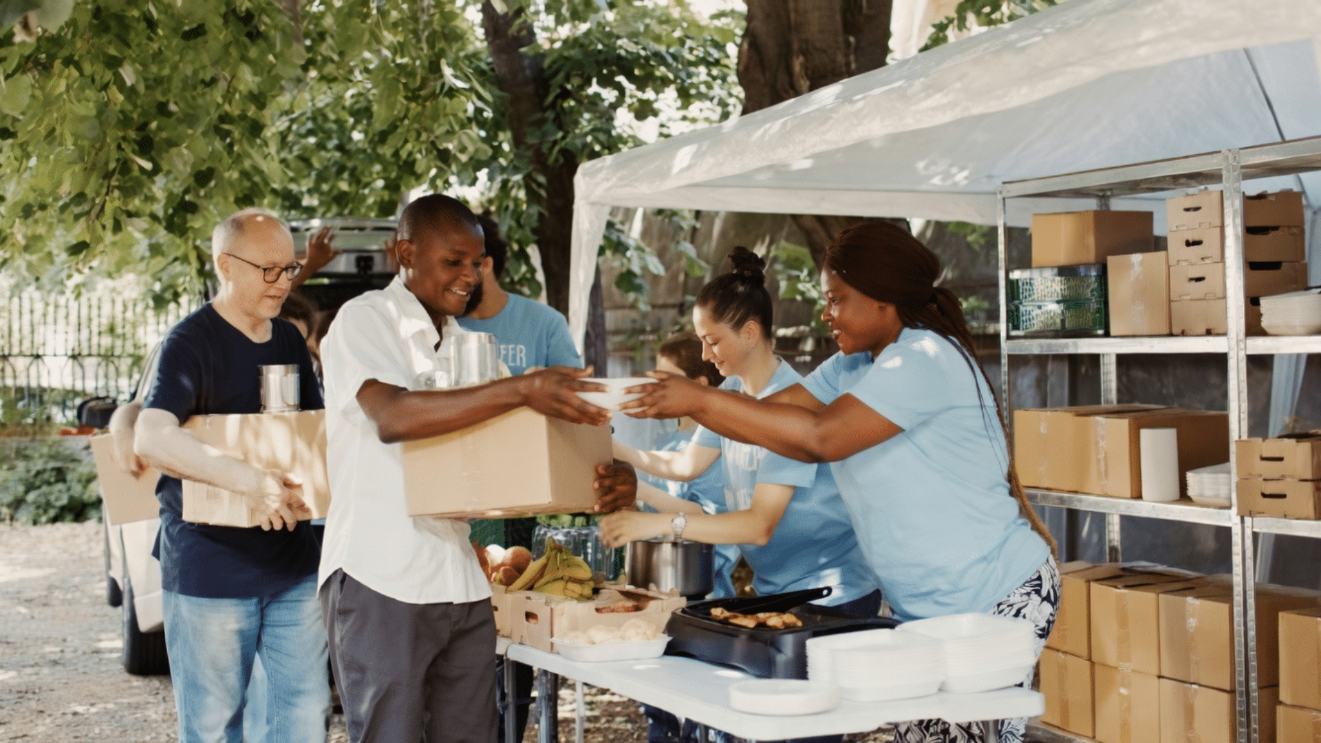 A diverse group of volunteers working outdoors at a community food bank, handing a container of food and a cardboard box of supplies to individuals in line.