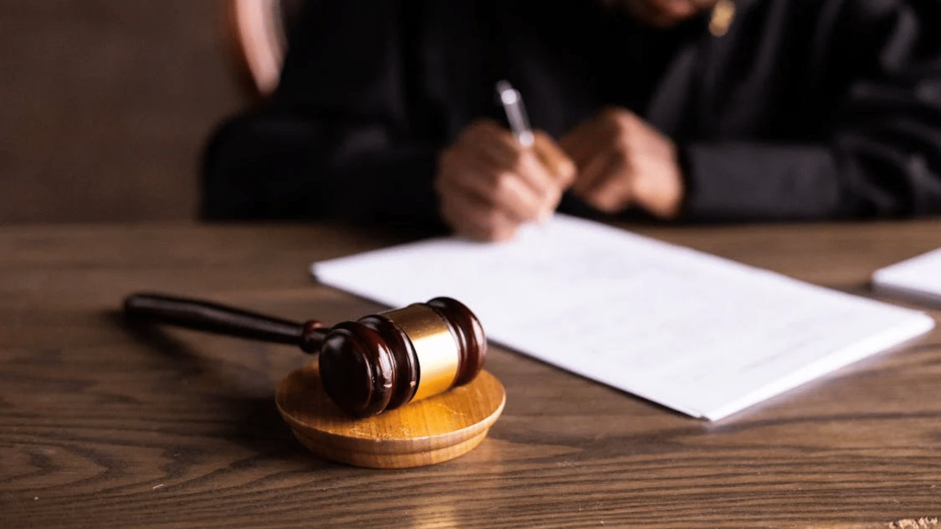 A wooden judge's gavel rests on a desk in front of a person writing on documents.