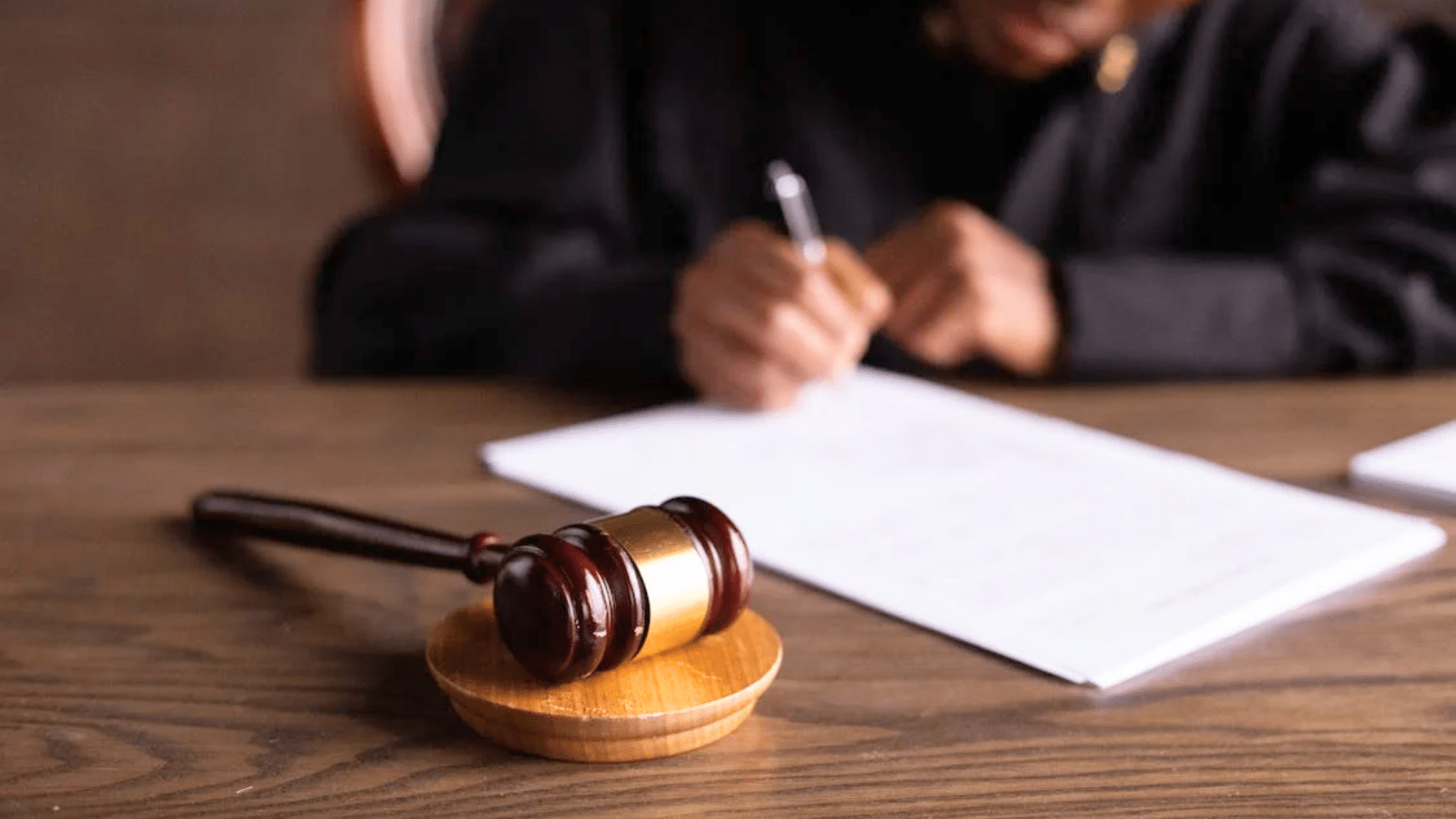 A wooden judge's gavel rests on a desk in front of a person writing on a document.