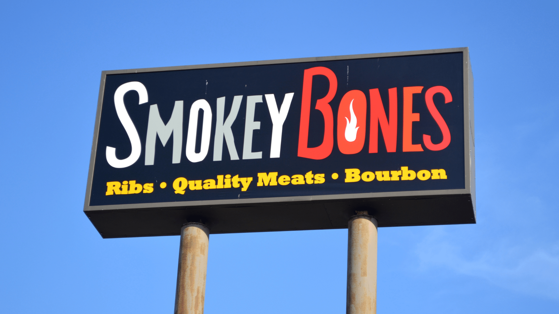 A large black sign for Smokey Bones raised on two poles against a bright blue sky.