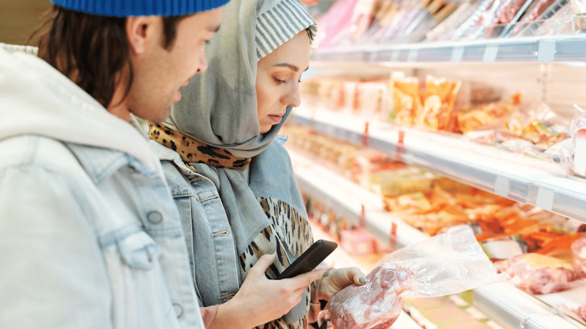 A man and a woman wearing a hijab look at a package of meat and a smartphone together in a grocery store aisle.