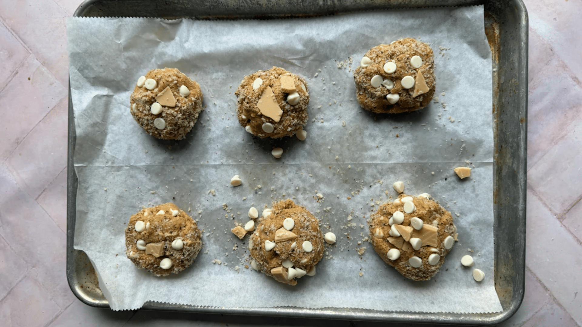 ix balls of cookie dough covered in white chocolate chips and butter chunks sitting on a baking sheet.