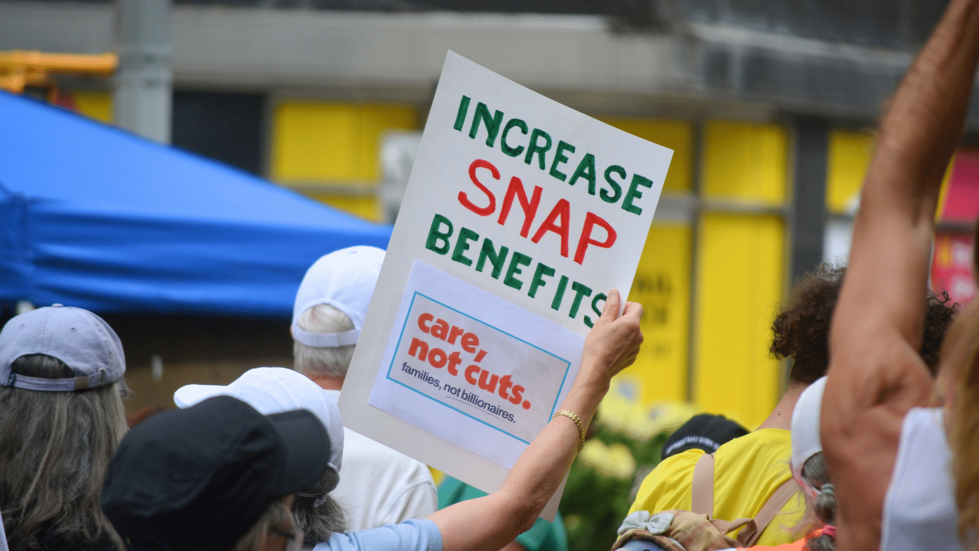 A person in a crowd holds up a protest sign that reads "INCREASE SNAP BENEFITS" and "care, not cuts."