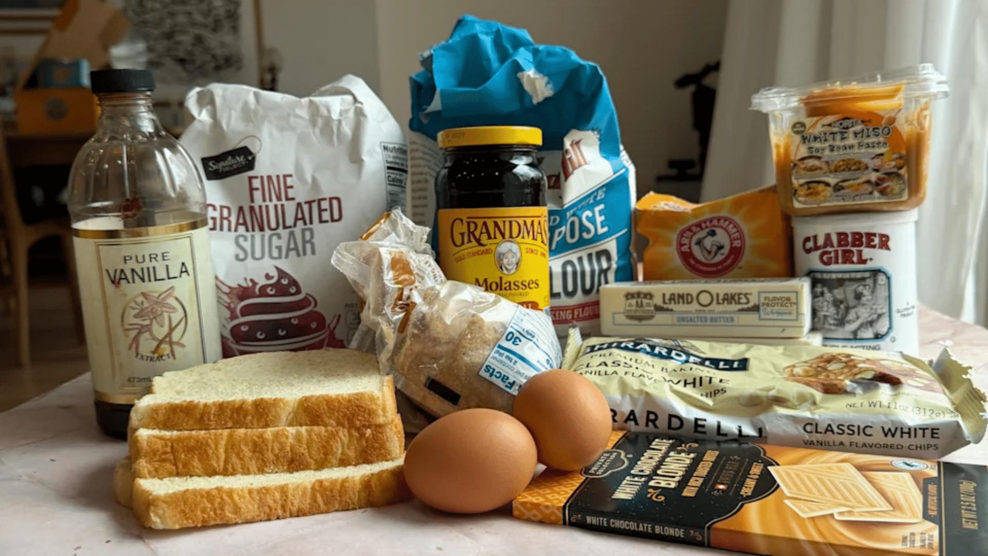 A collection of baking ingredients like flour, sugar, eggs, sliced bread, and chocolate chips gathered on a counter.