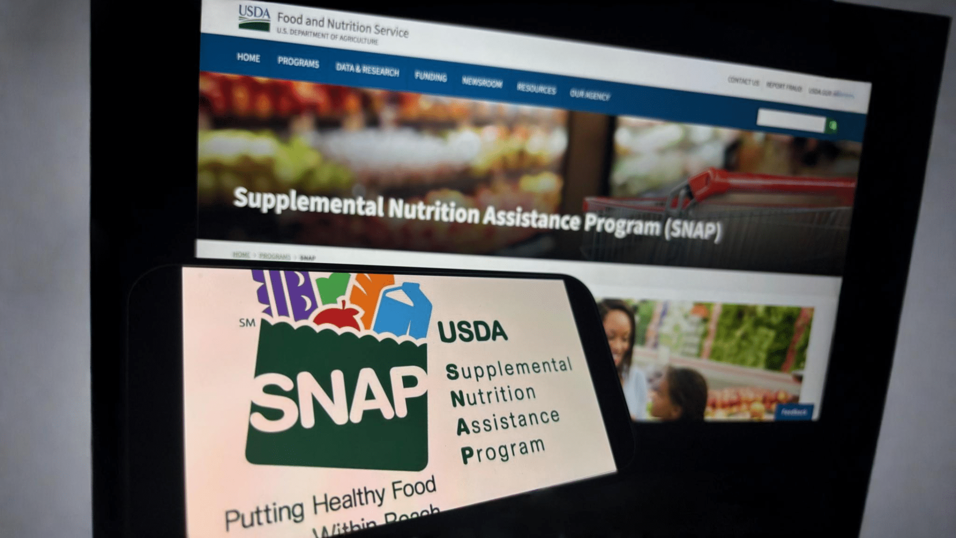 A smartphone displaying the USDA SNAP logo is held up in front of a computer monitor showing the program's official website.