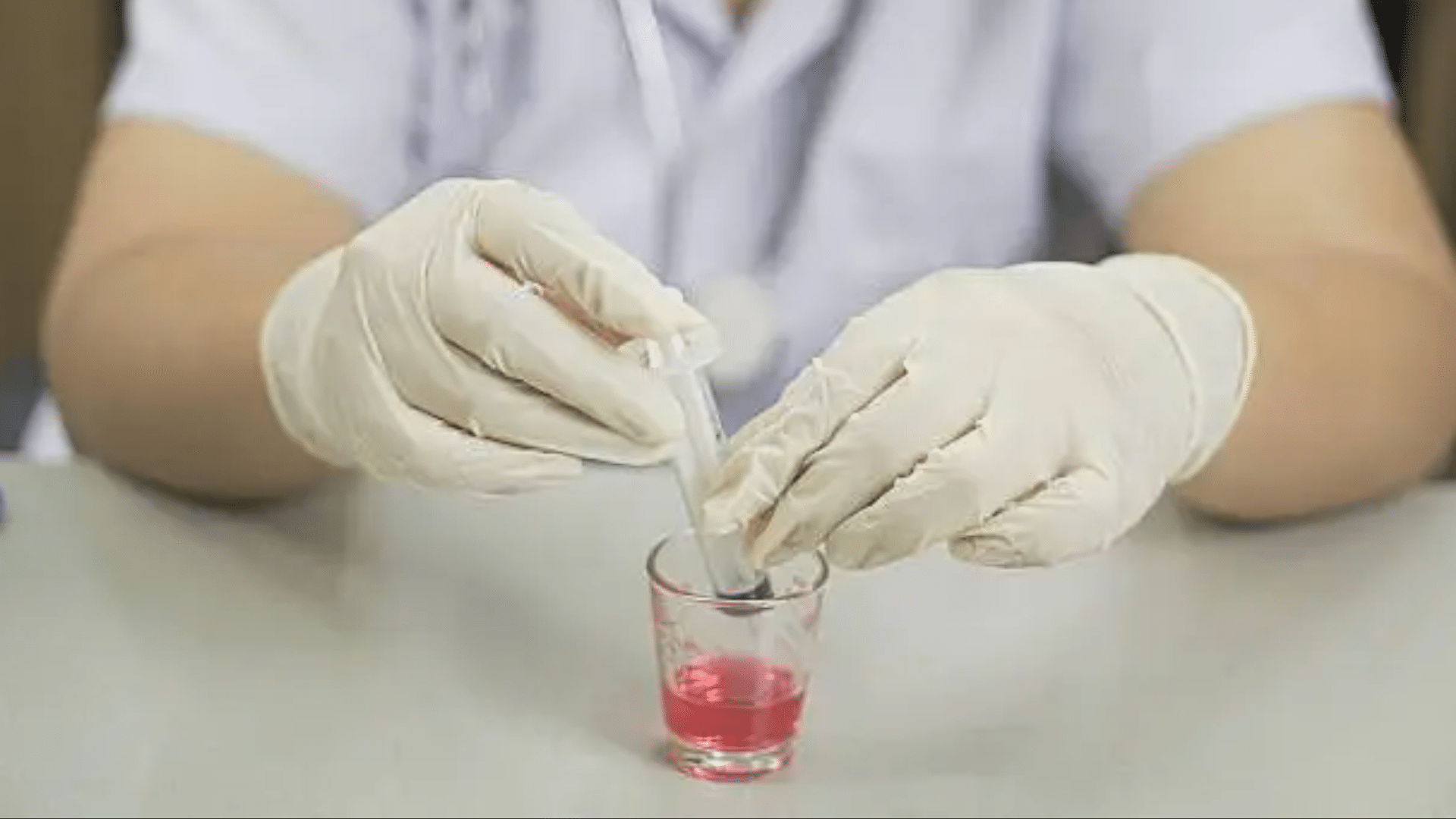 A close-up of a healthcare professional’s hands in white gloves using a syringe to draw a clear liquid from a small glass containing a bright pink liquid, representing laboratory testing or medication compounding.